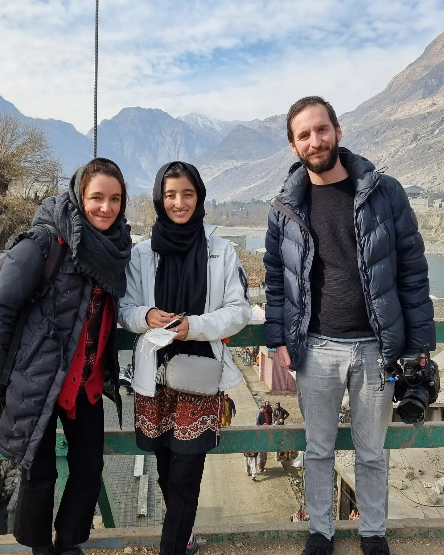 Three people standing outdoors with mountains in the background. Two women and one man, all dressed in jackets, smiling at the camera.