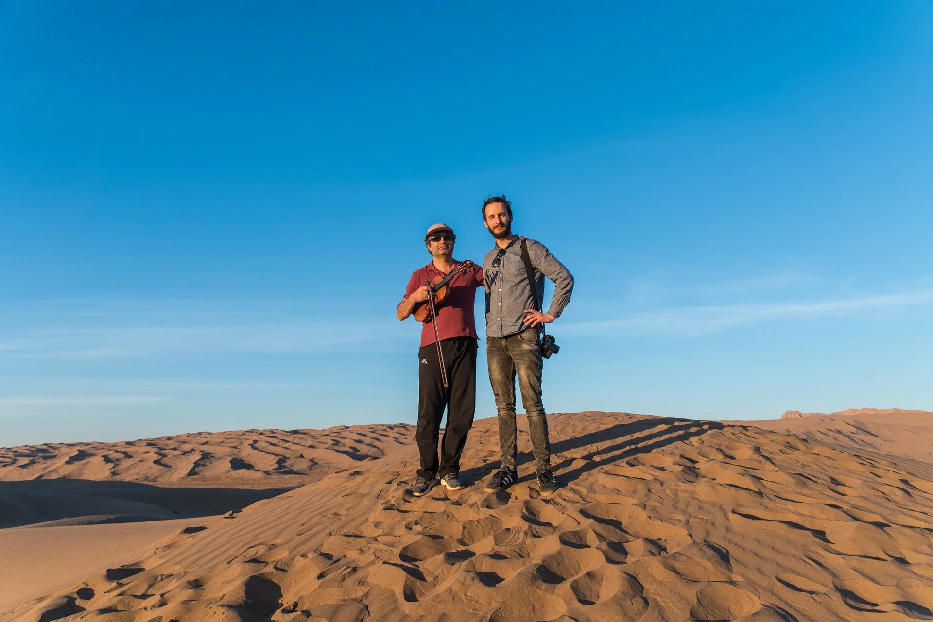Two men standing in desert sand dunes under a clear blue sky.