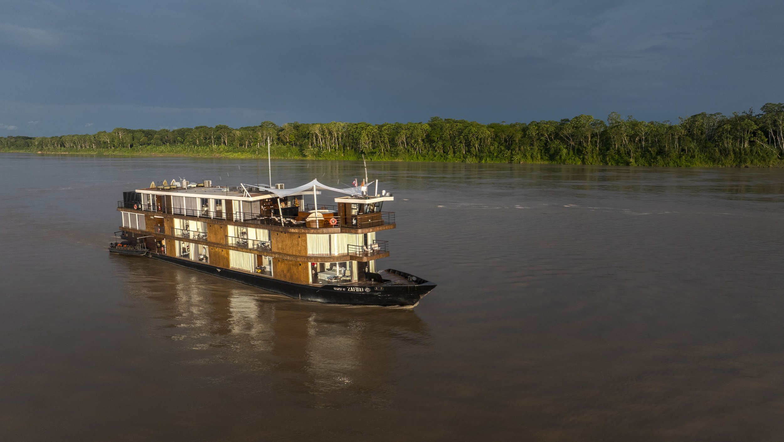 A riverboat cruising on a wide river with a green forested shoreline and a dark cloudy sky in the background.