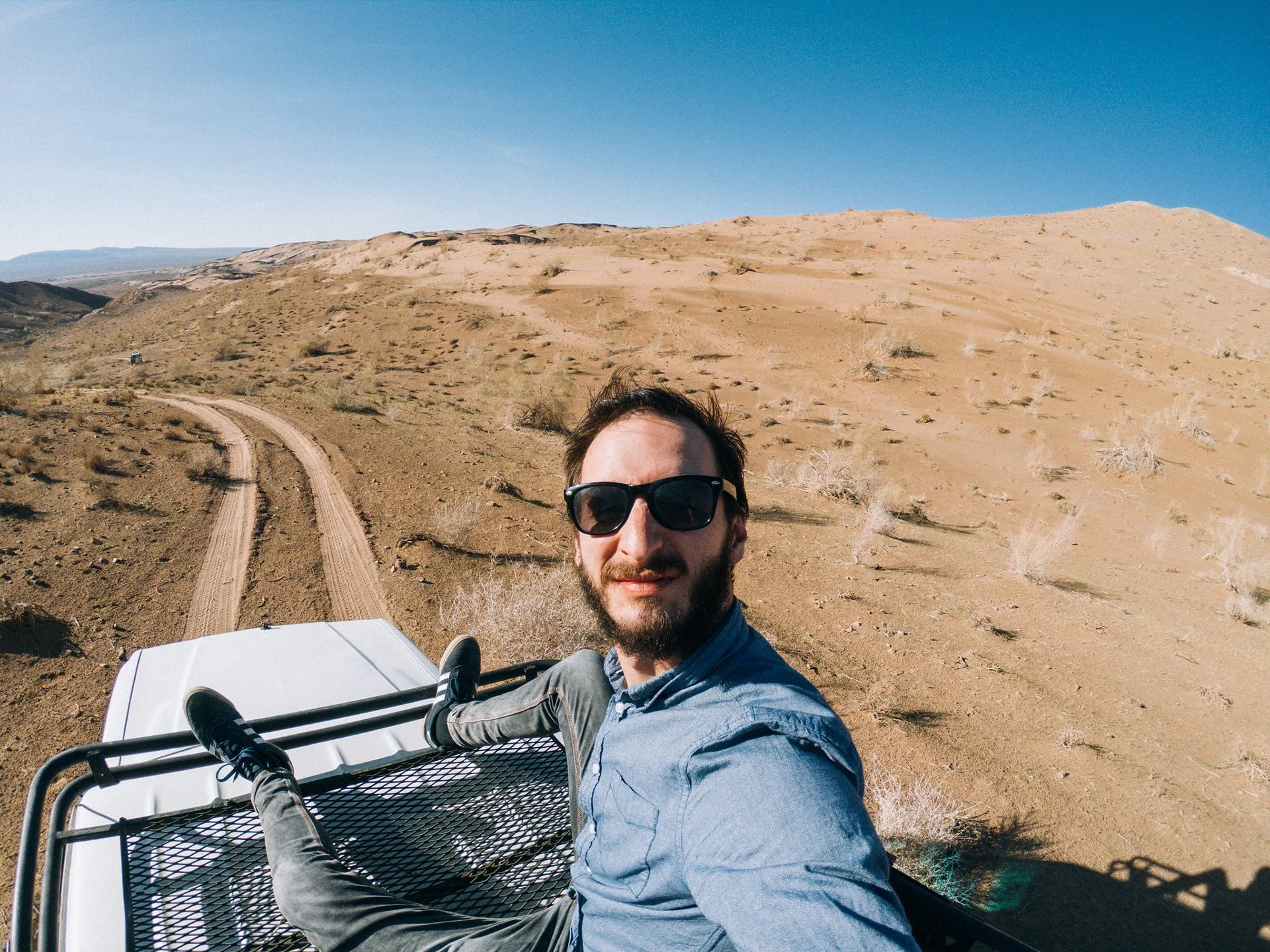 A man with dark hair and beard wearing sunglasses and a light blue shirt, sitting in a white off-road vehicle in a desert landscape with sandy hills and a clear blue sky.
