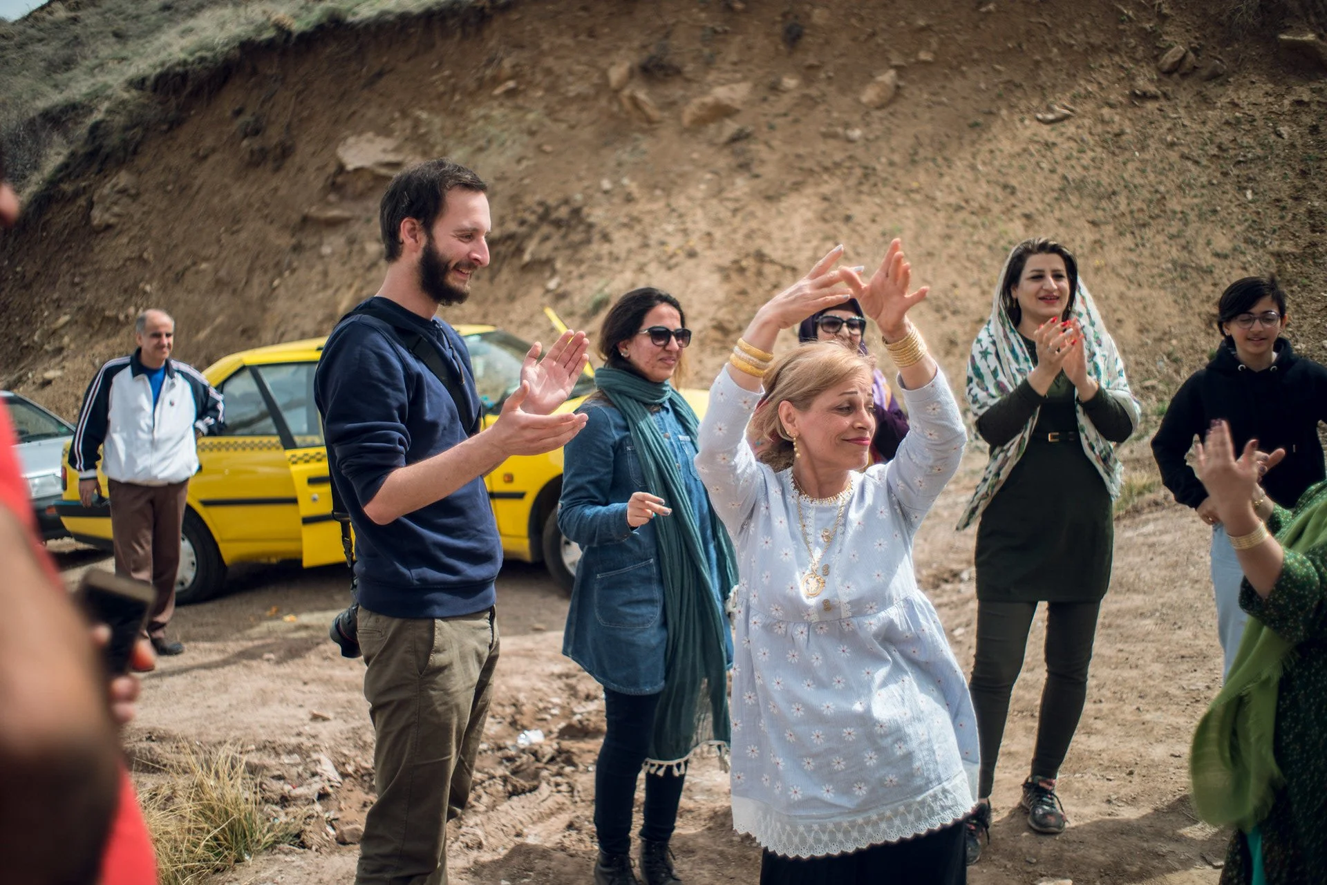 Group of diverse people outdoors, clapping and smiling during a gathering or celebration near a dirt hill, with cars parked in the background.