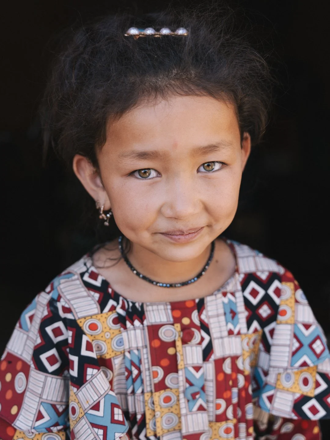 Close-up portrait of a young girl with long dark hair, hazel eyes, and light brown skin, wearing a colorful patterned dress, jewelry, and a small smile.