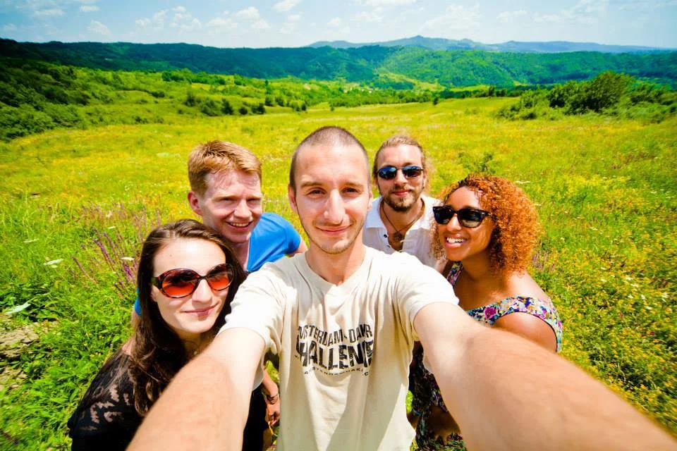 Group of five friends taking a selfie outdoors in a lush green field with mountains in the background.