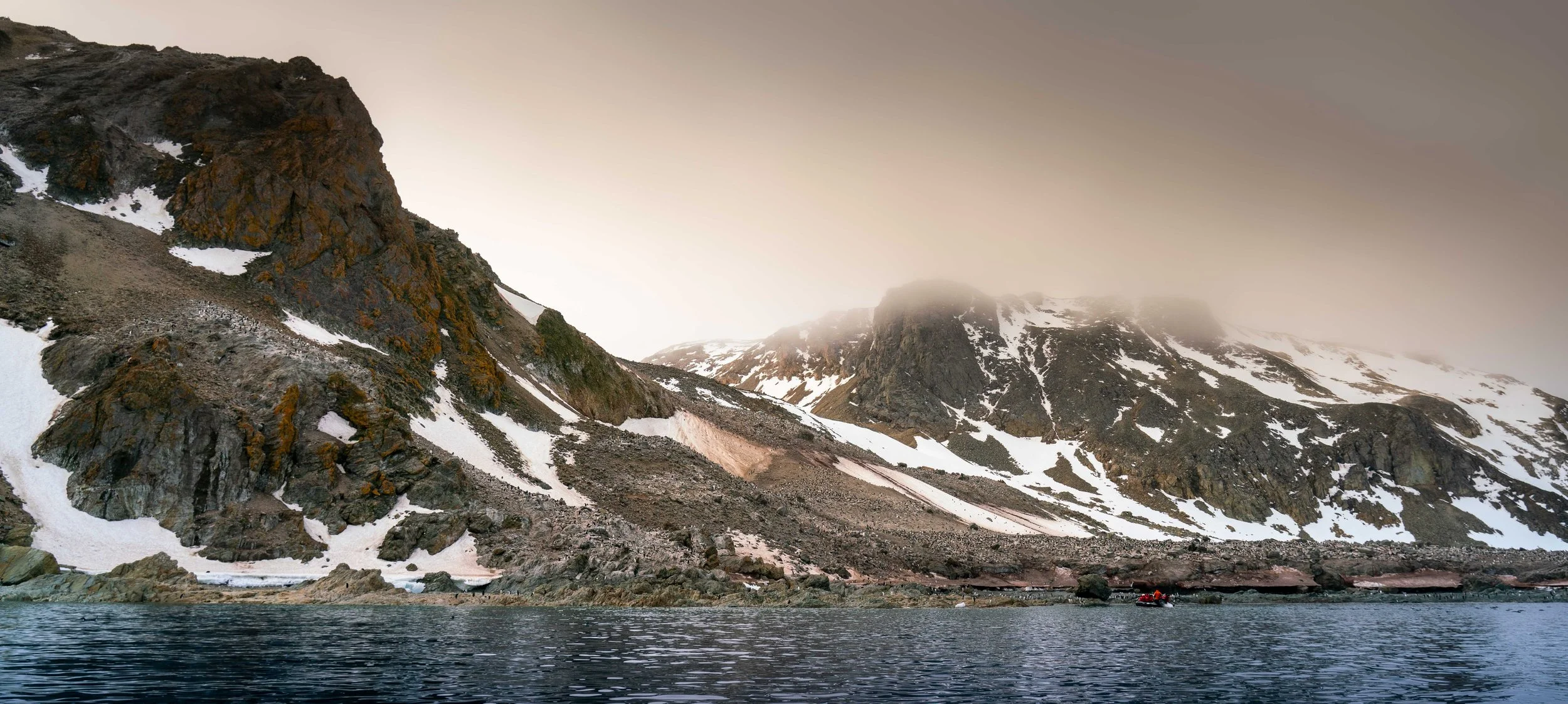 Mountain landscape with snow patches, cloudy sky, and a body of water with small boat in the foreground.
