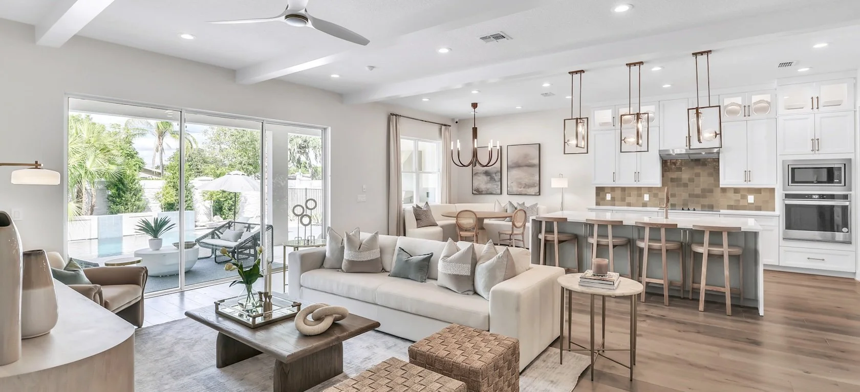 Bright living room and kitchen with white walls, sliding glass doors leading to outdoor patio, white sofa with multiple pillows, wooden coffee table, dining nook with chairs, white kitchen cabinets, island with bar stools, pendant lights, and modern appliances.