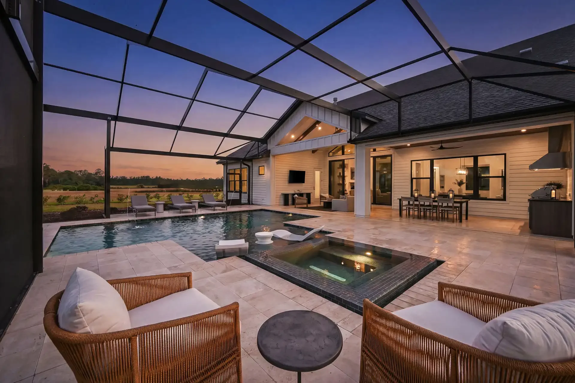 A backyard with a pool and hot tub on a patio under a glass-covered enclosure at sunset. Lounge chairs and outdoor furniture are visible, with a view of open land and trees in the background.