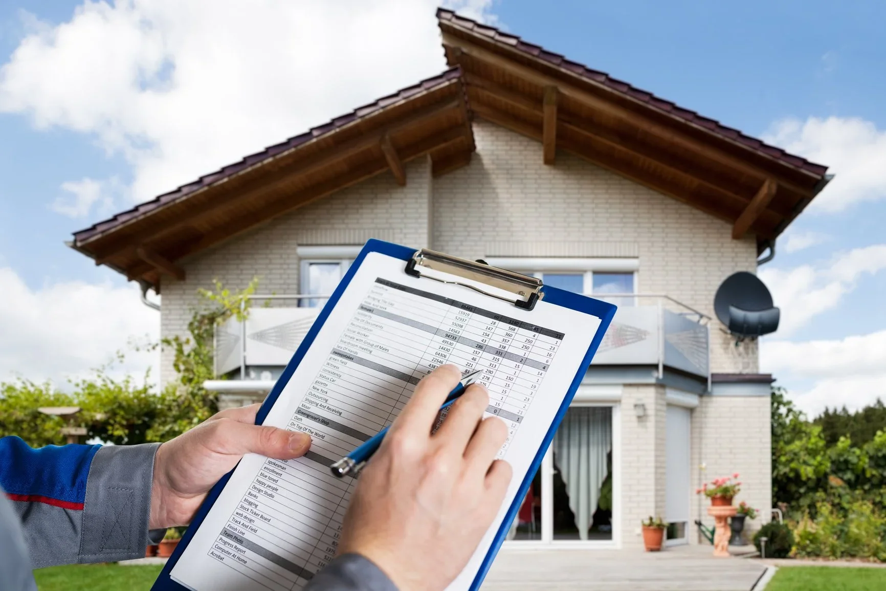 Person holding a clipboard with a spreadsheet outside a house with a brick facade, balcony, satellite dish, and potted plants.