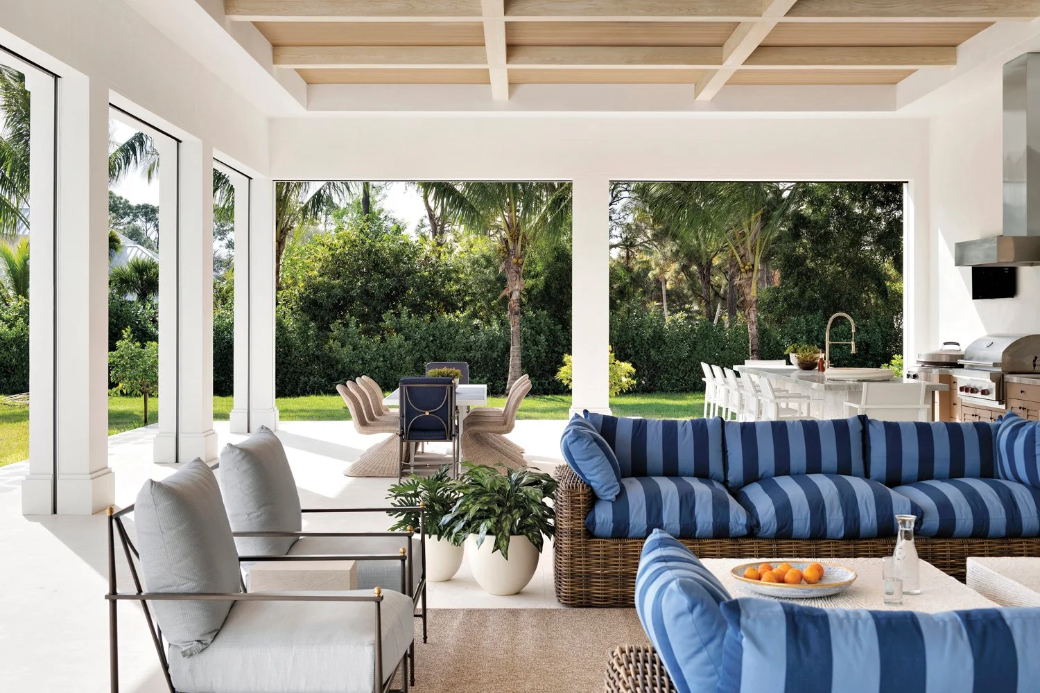 Indoor-outdoor living space with white walls, ceiling, and trim; wicker and metal chairs with white cushions; blue-striped outdoor sofa; potted plants; view of lush green yard with palm trees; outdoor dining table with chairs; white kitchen island with barstools and outdoor grill.