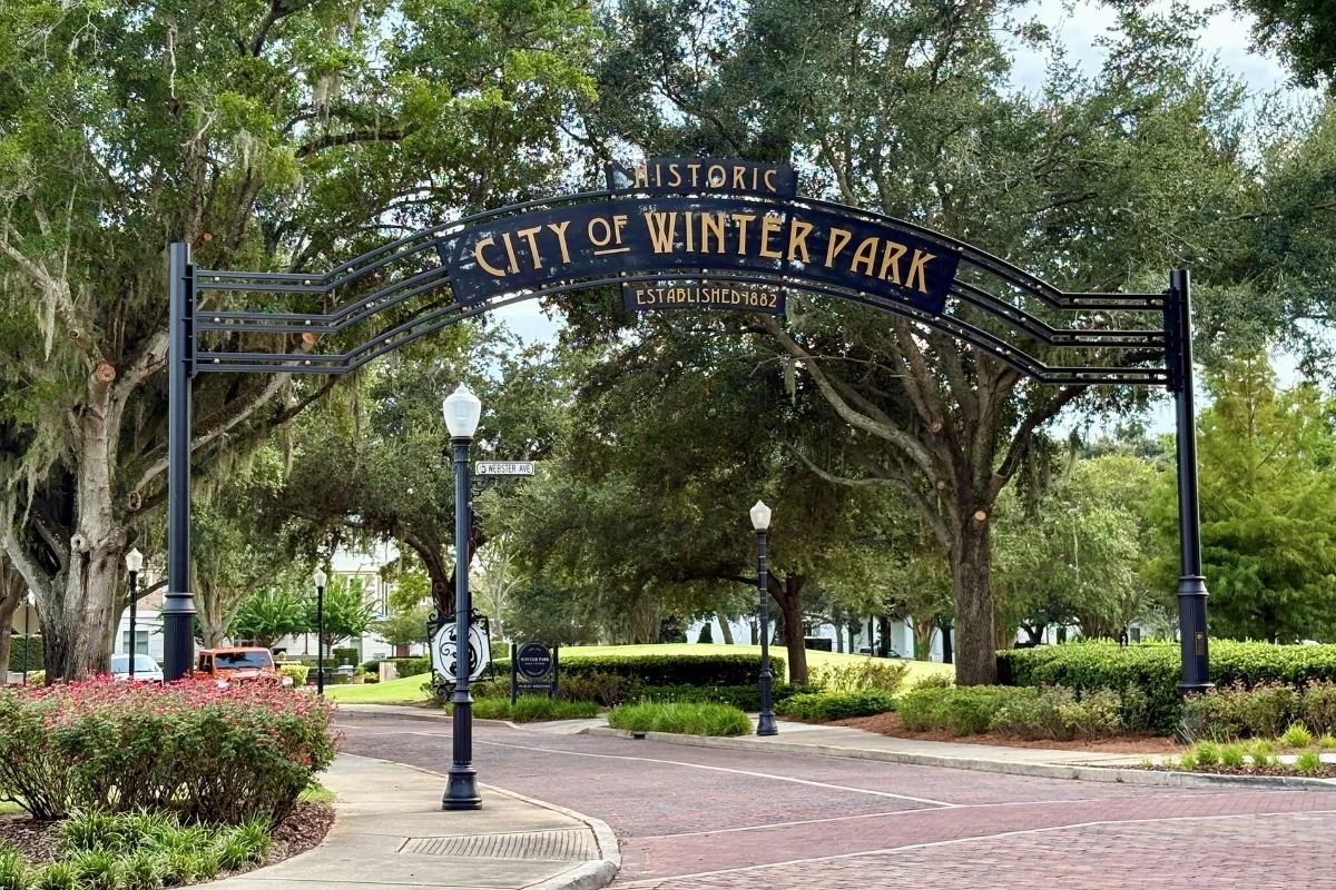 Entrance to Winter Park, featuring a decorative black arch sign reading 'Historic City of Winter Park, Established 1882,' with trees, street lamps, and a brick-paved street in the background.