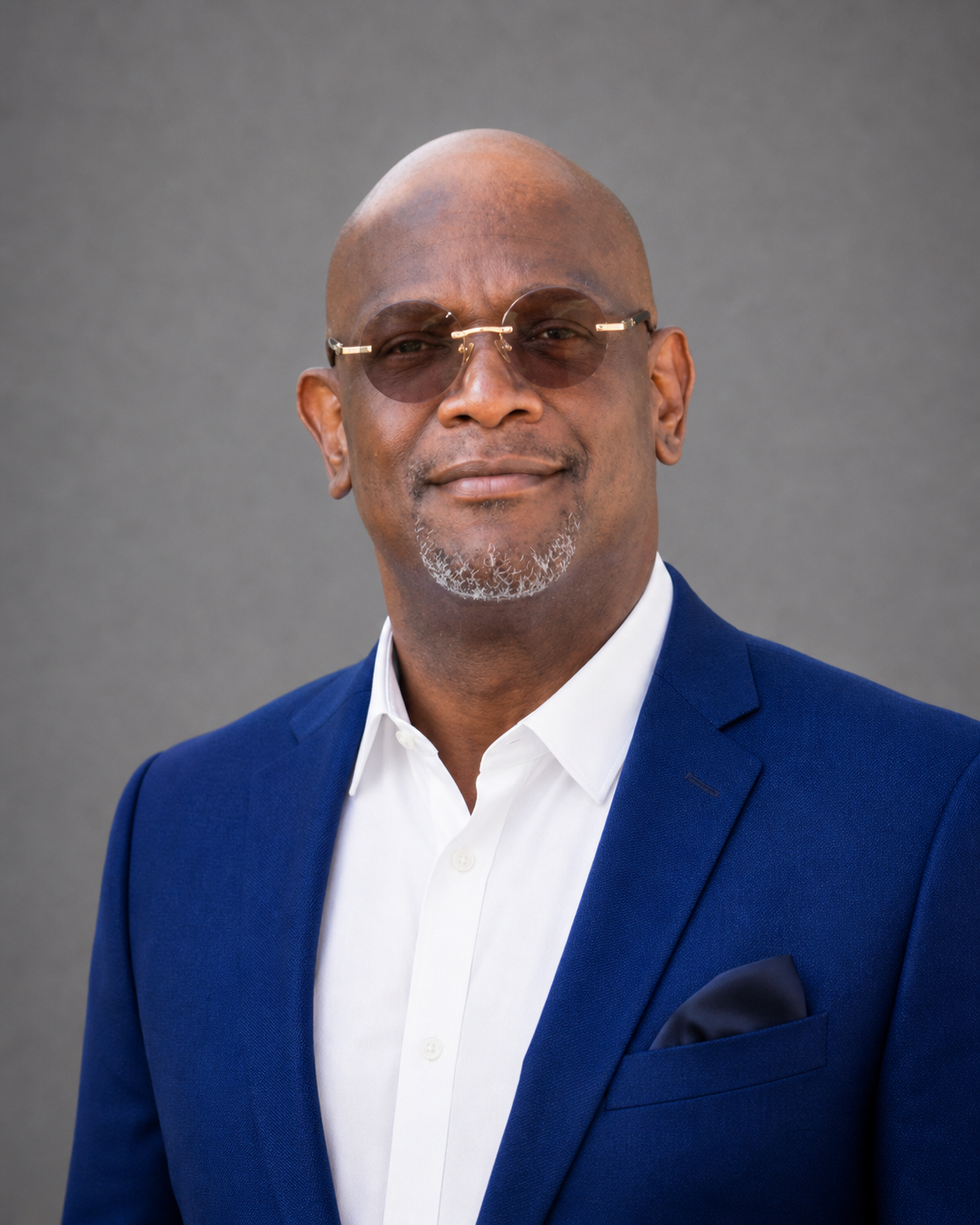 A man wearing a blue suit, white shirt, sunglasses, and a pocket square. Gray background.