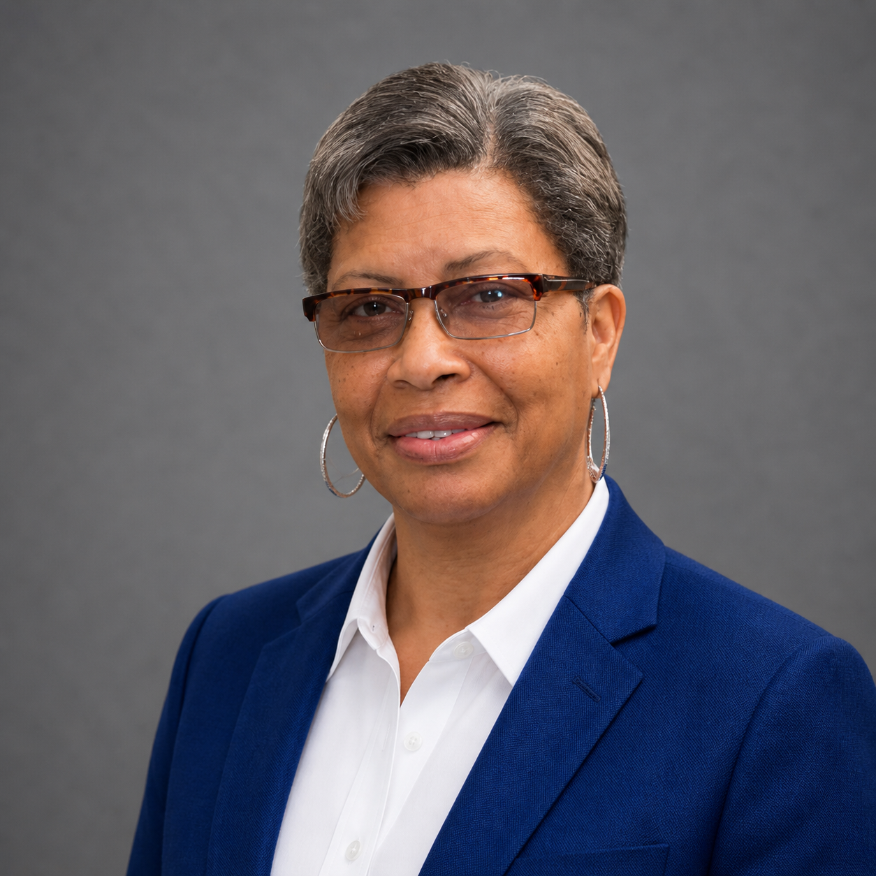 A professional woman with short gray hair, wearing glasses, hoop earrings, a white shirt, and a blue blazer, standing against a gray background.
