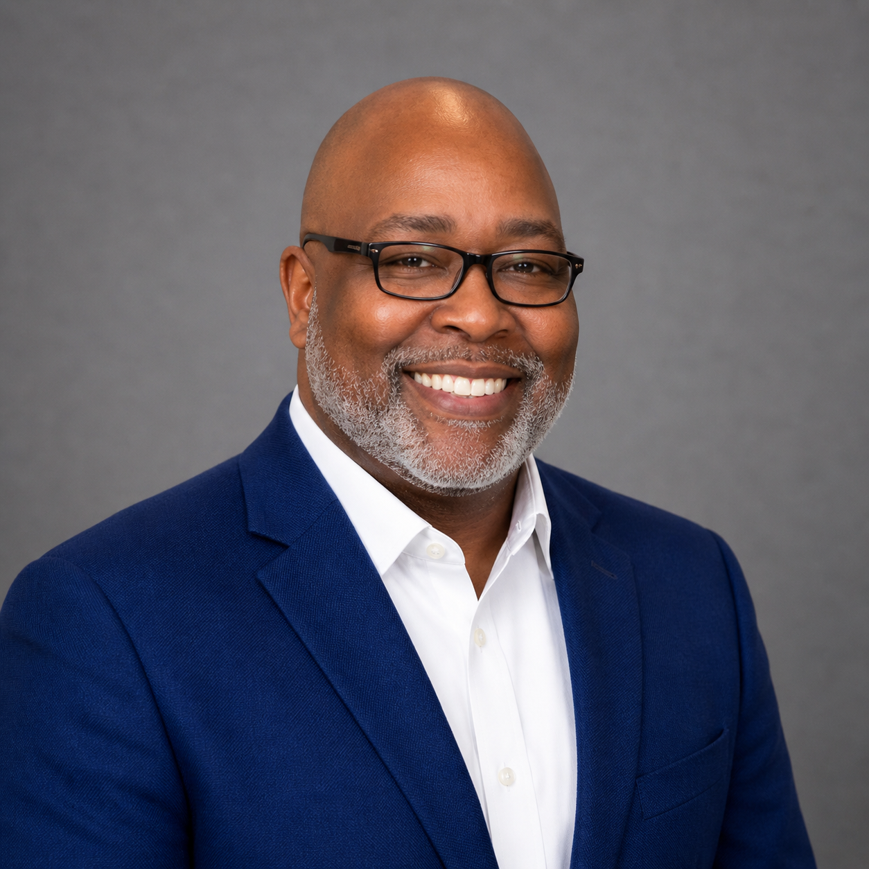 A smiling African American man with glasses, a white beard, dressed in a navy blue blazer and white shirt, against a gray background.