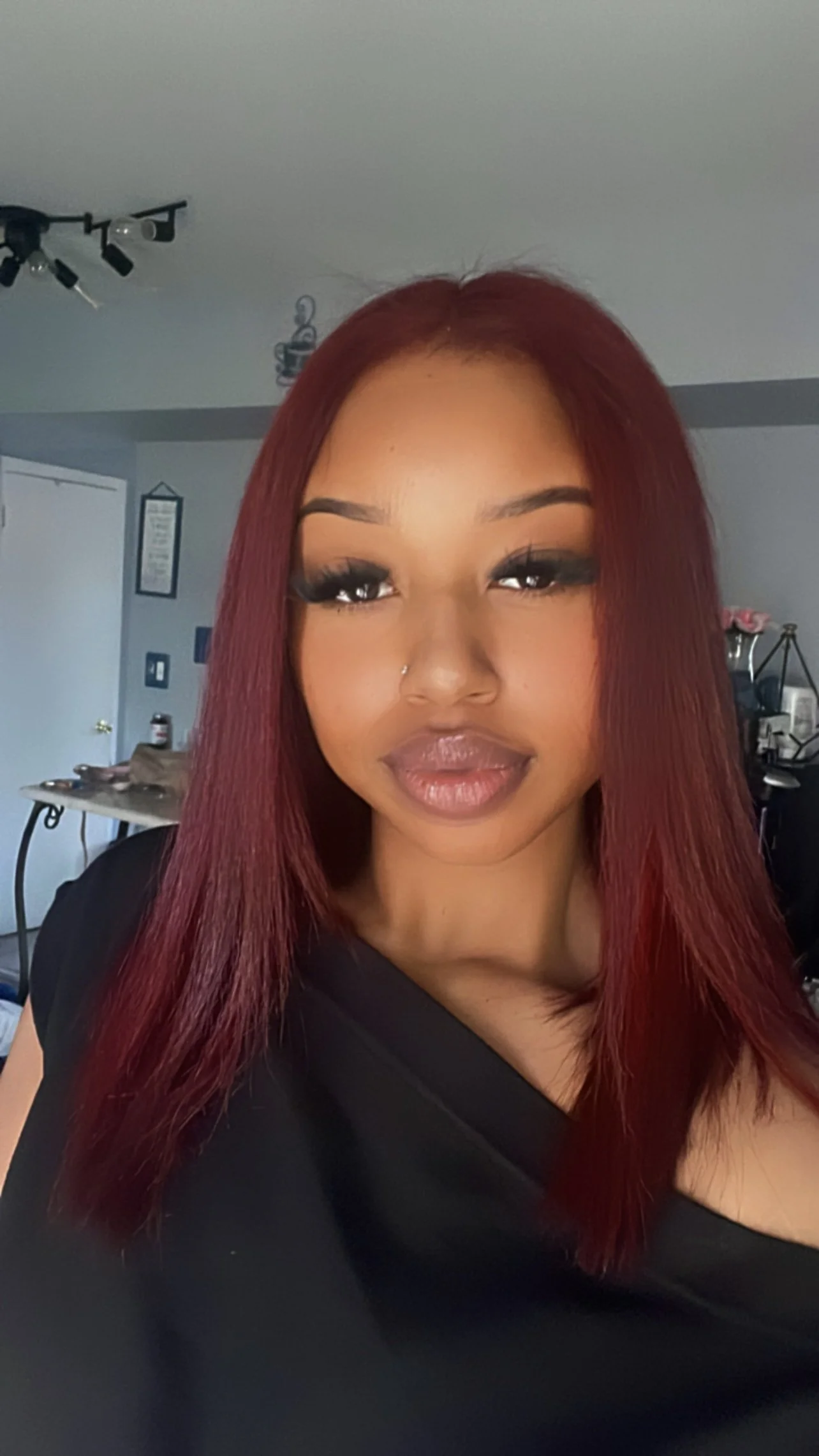 A woman with long red hair, wearing makeup including false eyelashes and lipstick, poses for a selfie in a room with a gray ceiling, a ceiling fan, and various household items in the background.