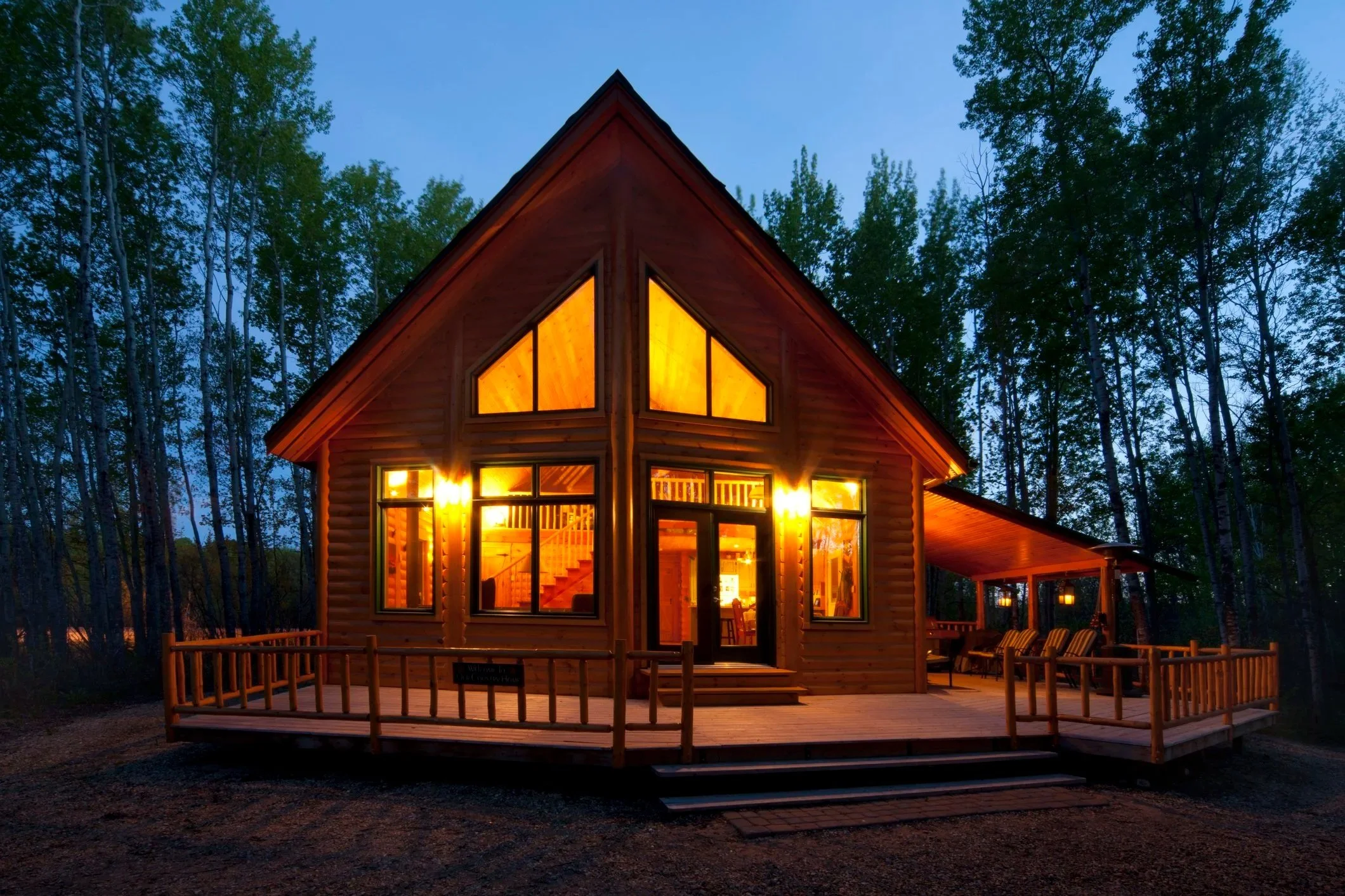 A cozy wooden house in a forest at dusk with large lit windows and a warm interior glow