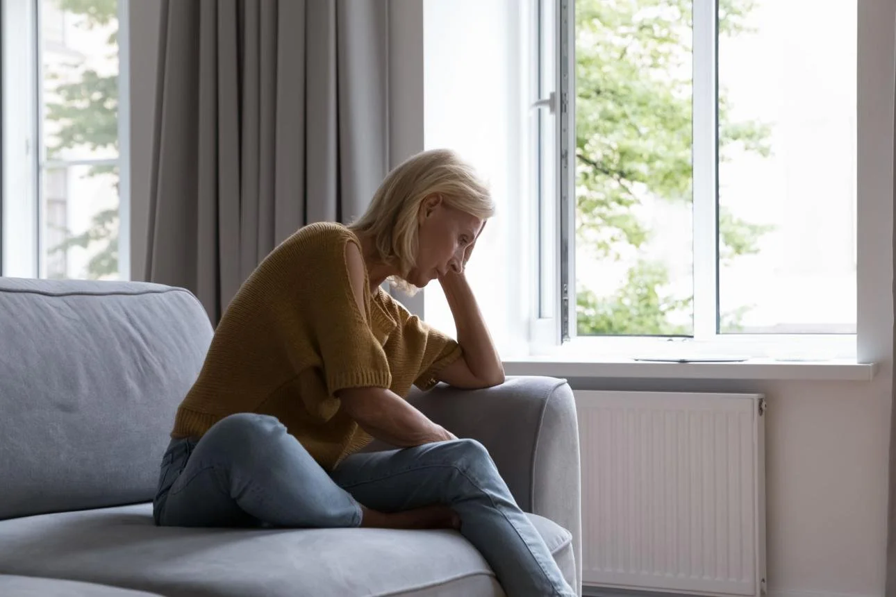 An elderly woman sitting on a light gray sofa near an open window, wearing a brown sweater and jeans, with a contemplative or sad expression.