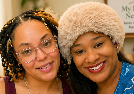 Two women smiling closely together, one wearing glasses and a decorative hairstyle, the other wearing a furry hat and earrings, in an indoor setting.