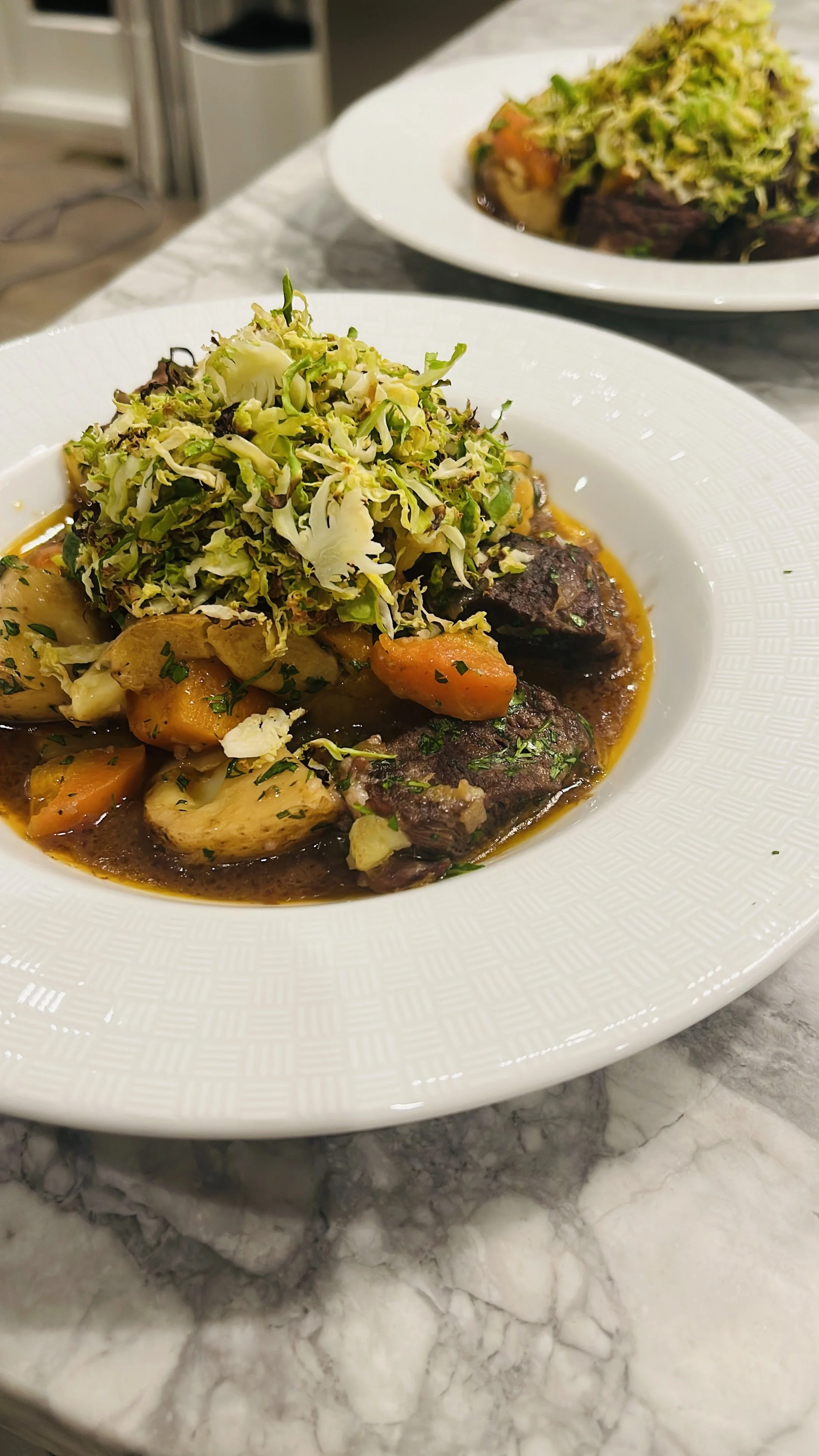 A white plate with beef stew, carrots, and potatoes topped with shredded Brussels sprouts on a marble countertop.