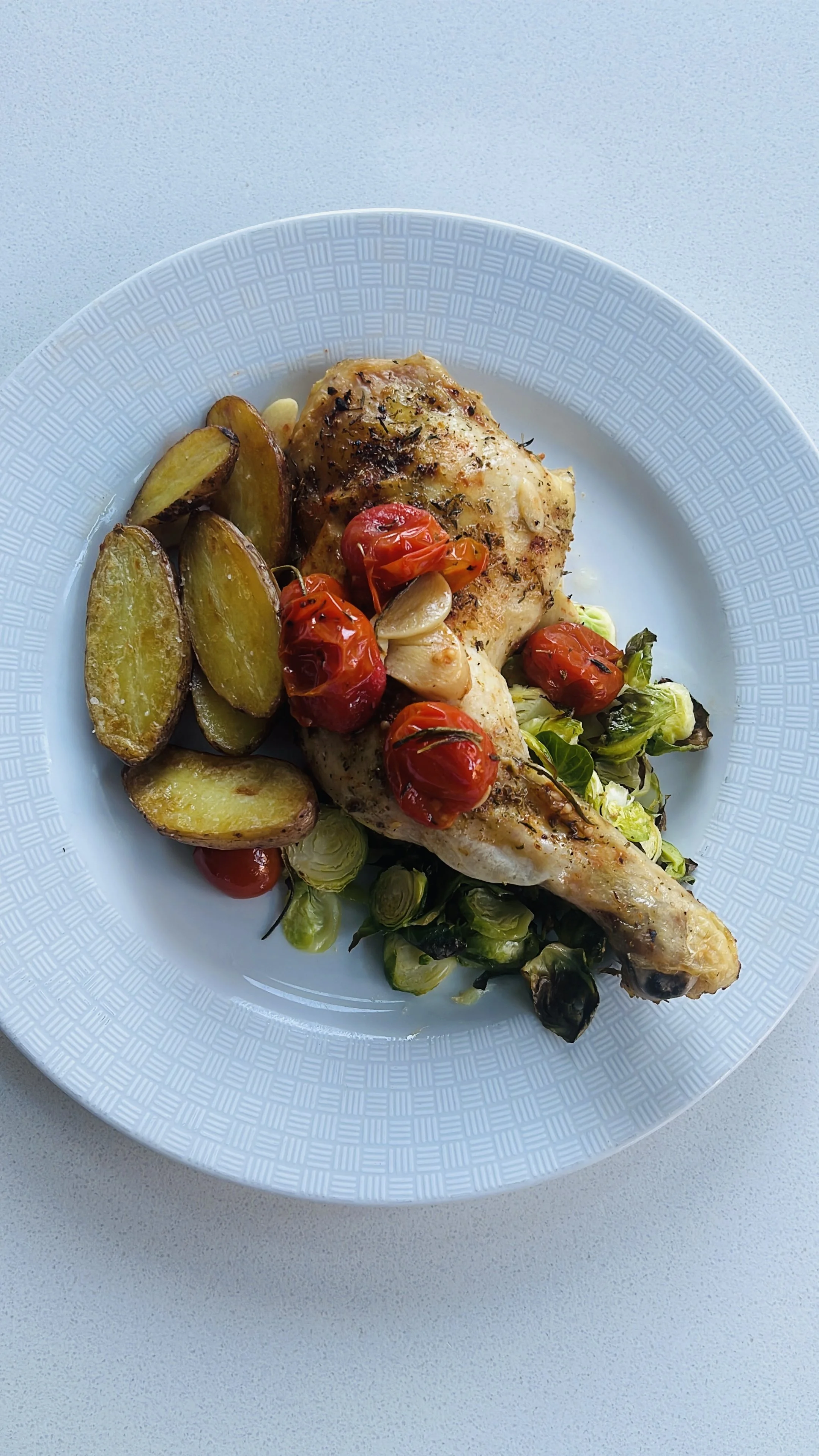 Roasted chicken leg with cherry tomatoes, garlic, and roasted potato wedges on a white plate.
