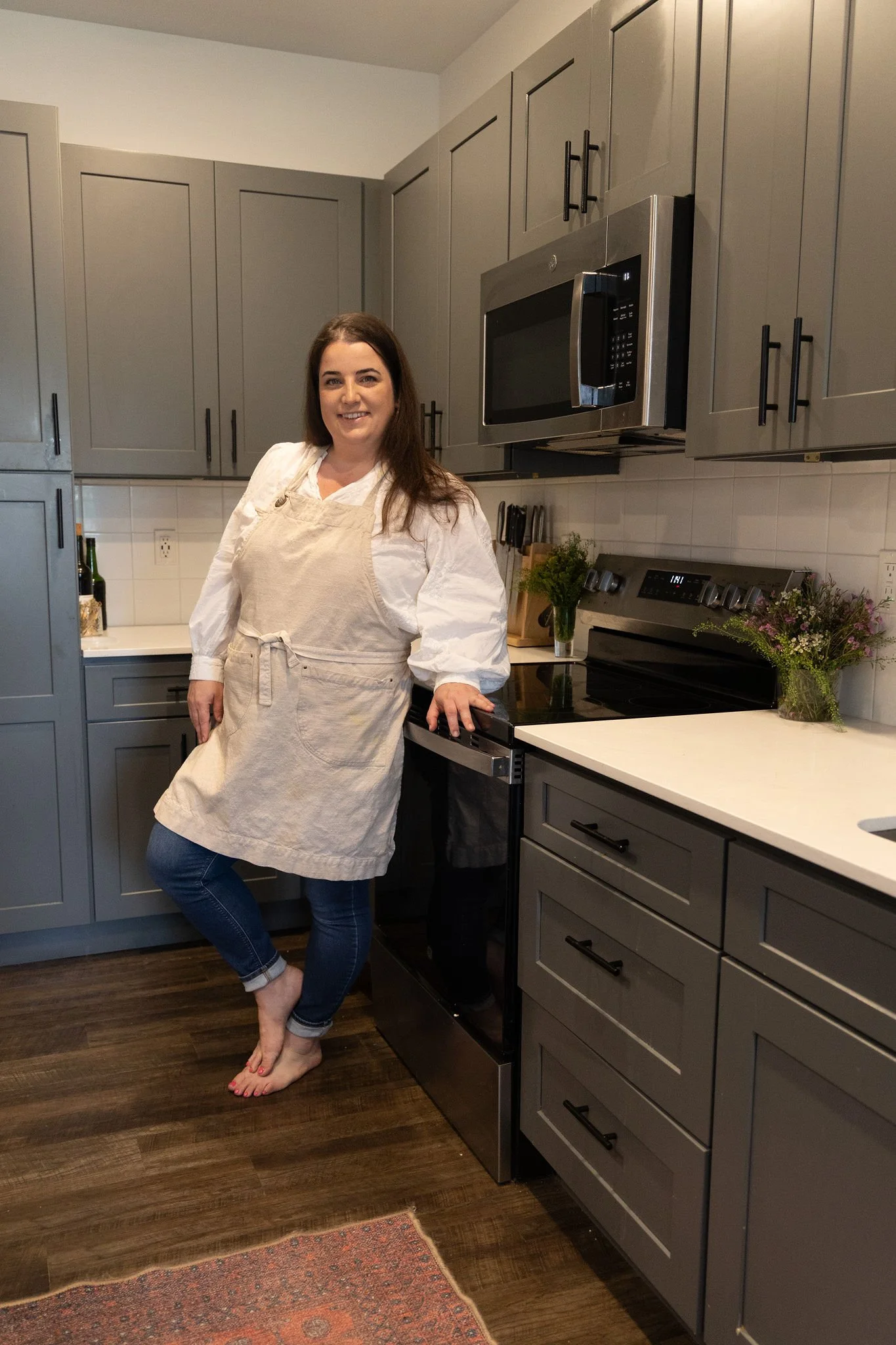 A woman standing barefoot in a modern kitchen, wearing a white shirt and beige apron, with dark hair. The kitchen has gray cabinets, dark wood flooring, and a stainless steel microwave and oven. There are flowers on the counter behind her.