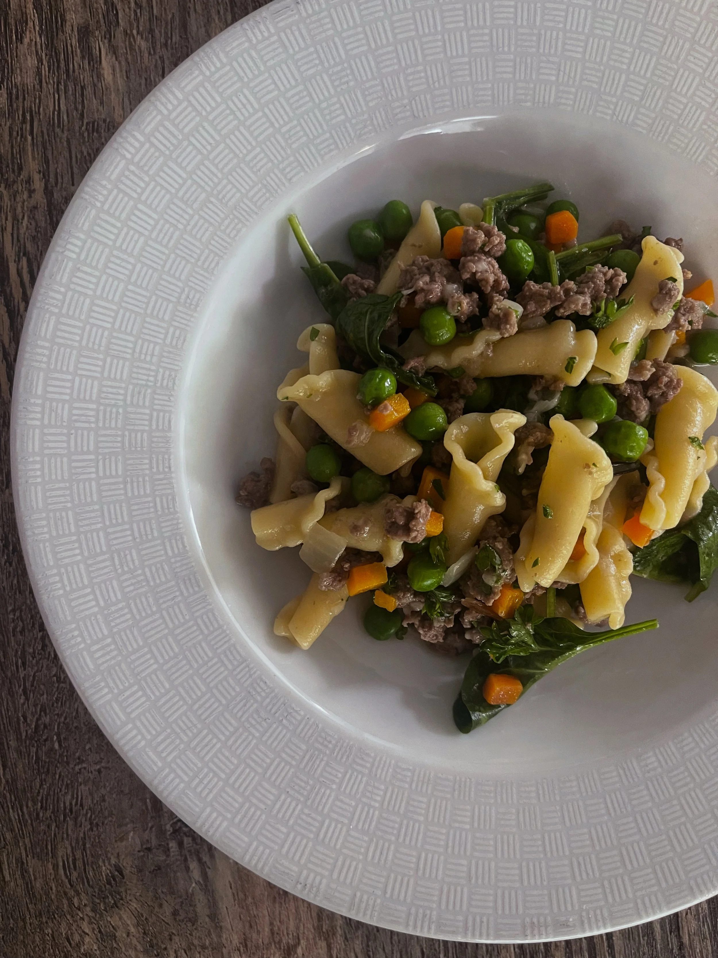 Plate of pasta with ground beef, peas, carrots, and spinach on a wooden table.