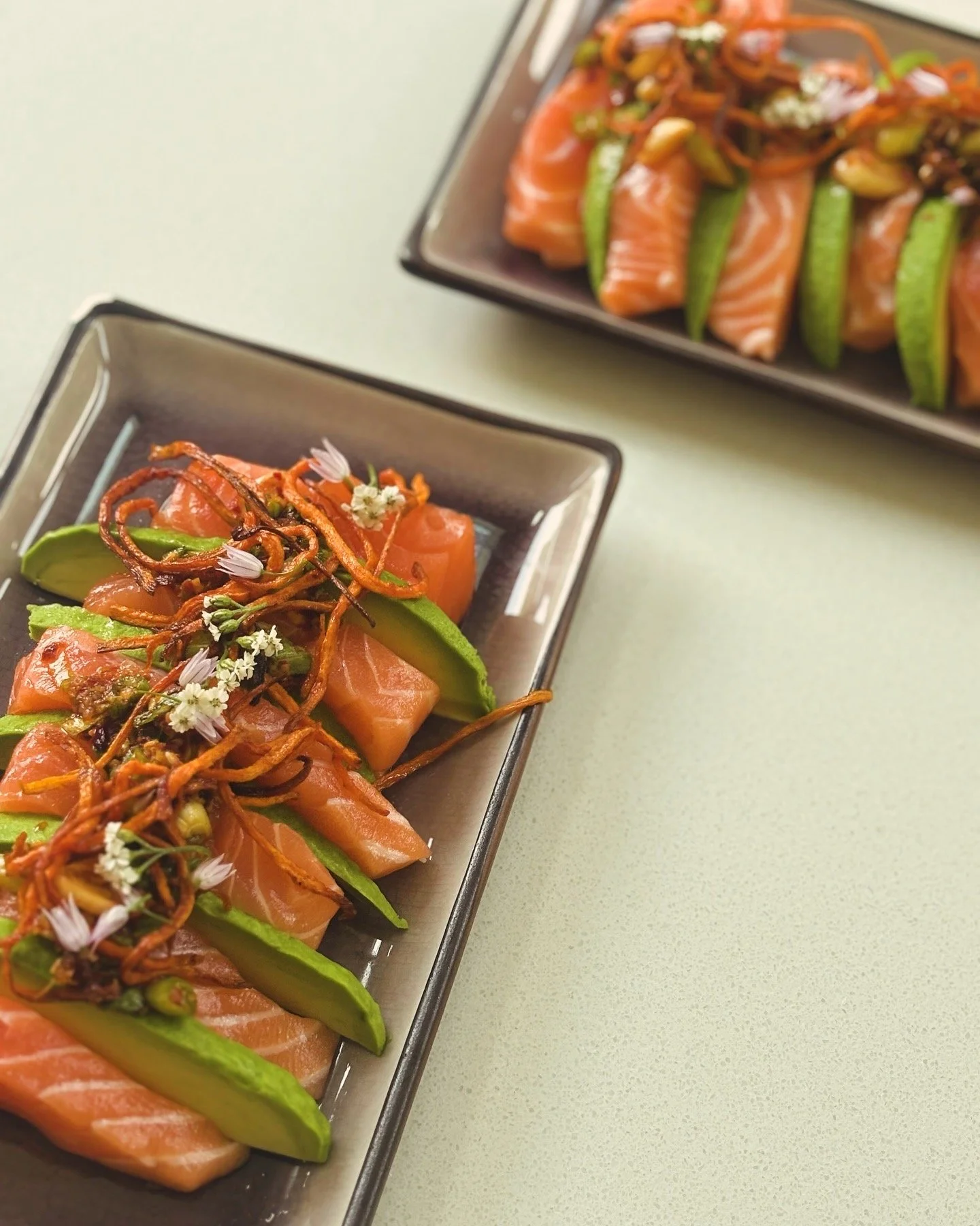 Fresh slices of salmon, green snap peas, and garnishes with edible flowers on a black rectangular plate.