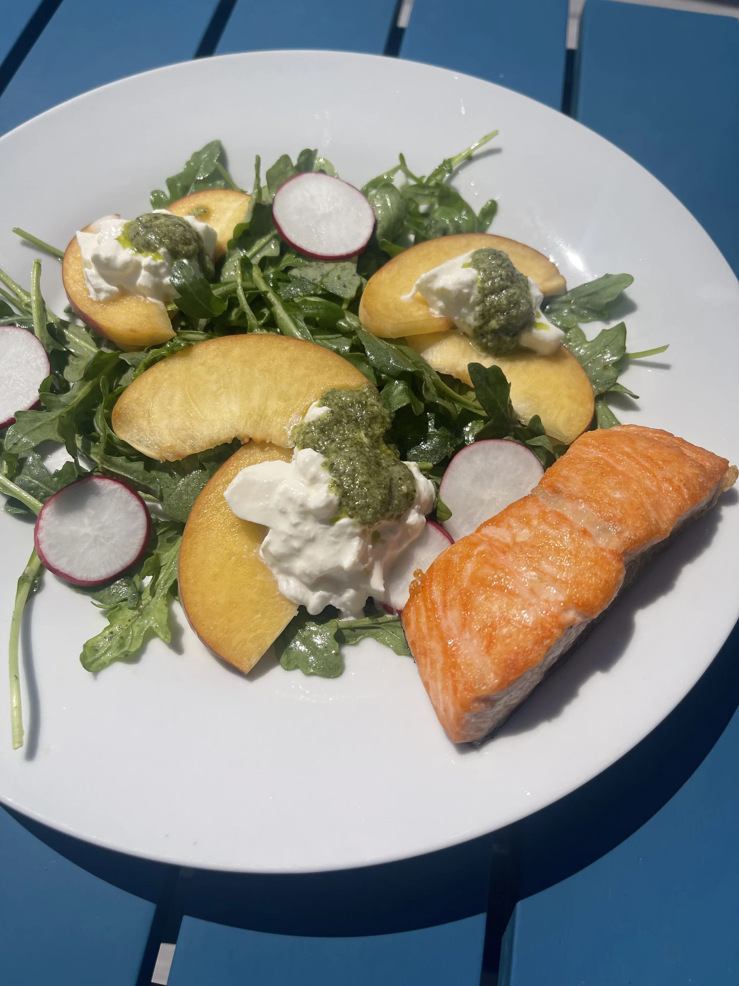 A white plate containing a salad with arugula, sliced radishes, peach slices with dollops of white cheese and green pesto, and a piece of cooked salmon. The plate is on a blue table outside in sunlight.