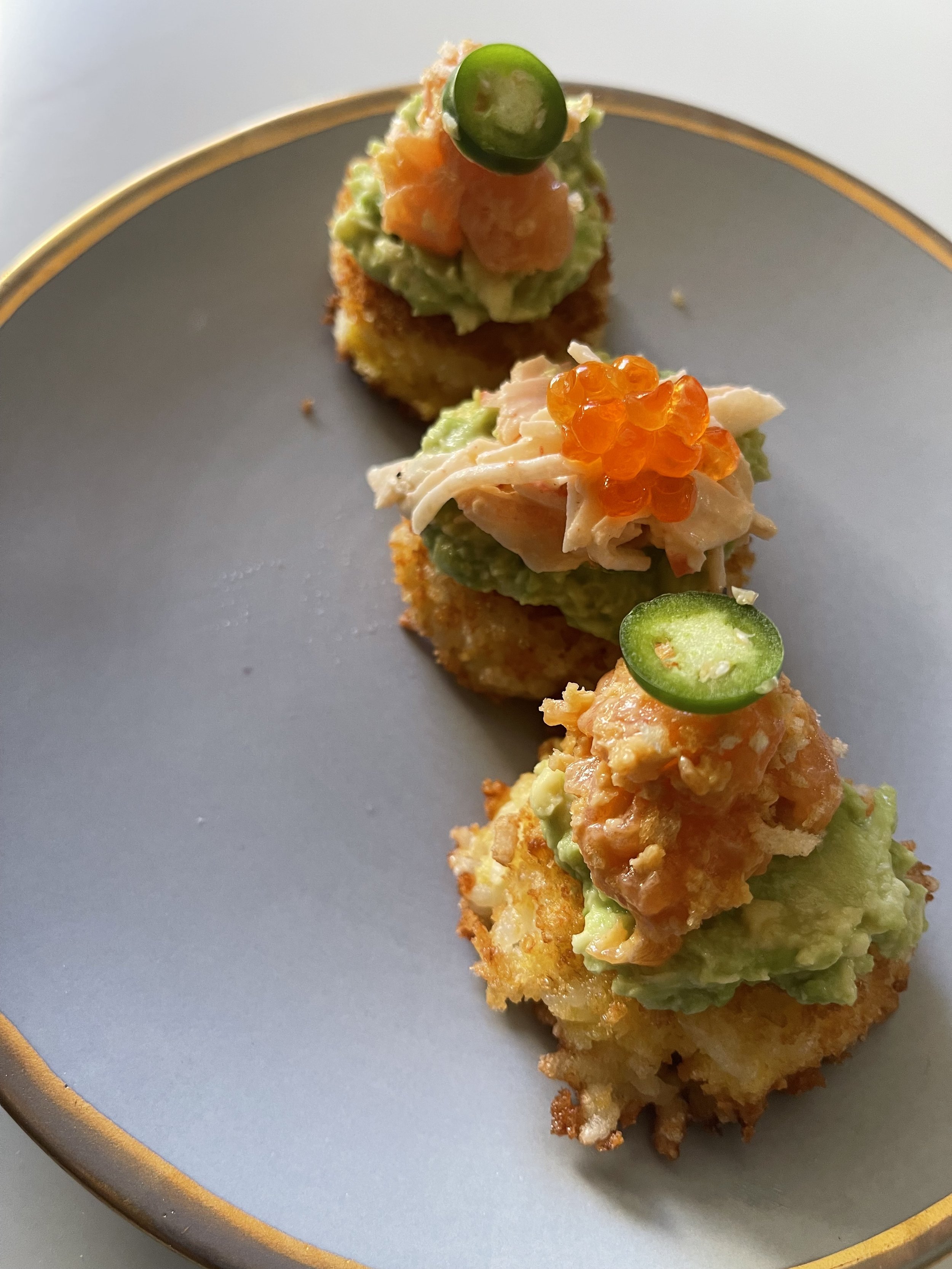 Close-up of three pieces of sushi topped with avocado, orange fish roe, and sliced jalapeño on a gray plate with a gold rim.
