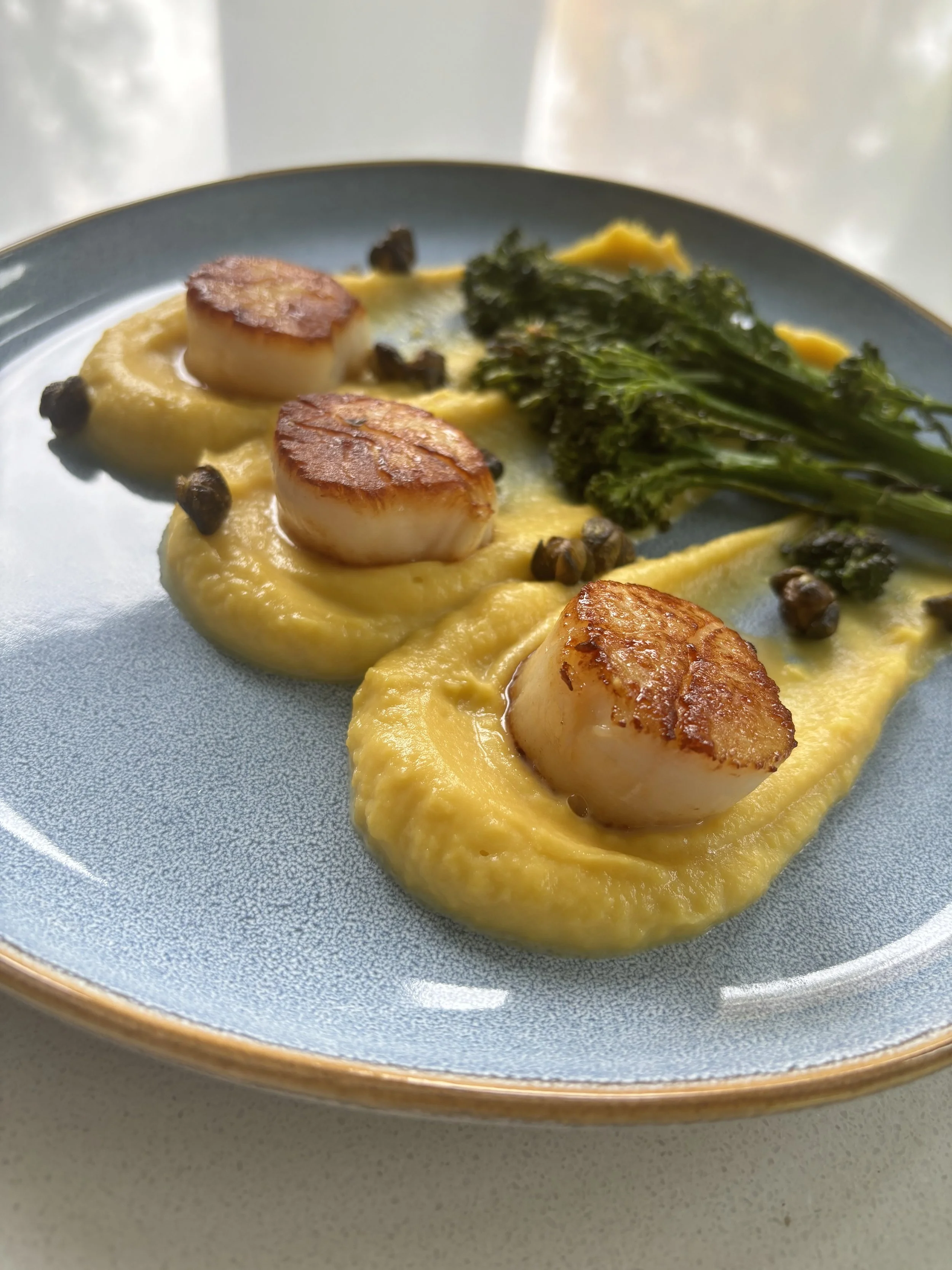 Seared scallops served on a bed of creamy yellow sauce, garnished with small black peppercorns and accompanied by a portion of sautéed kale on a light blue plate.