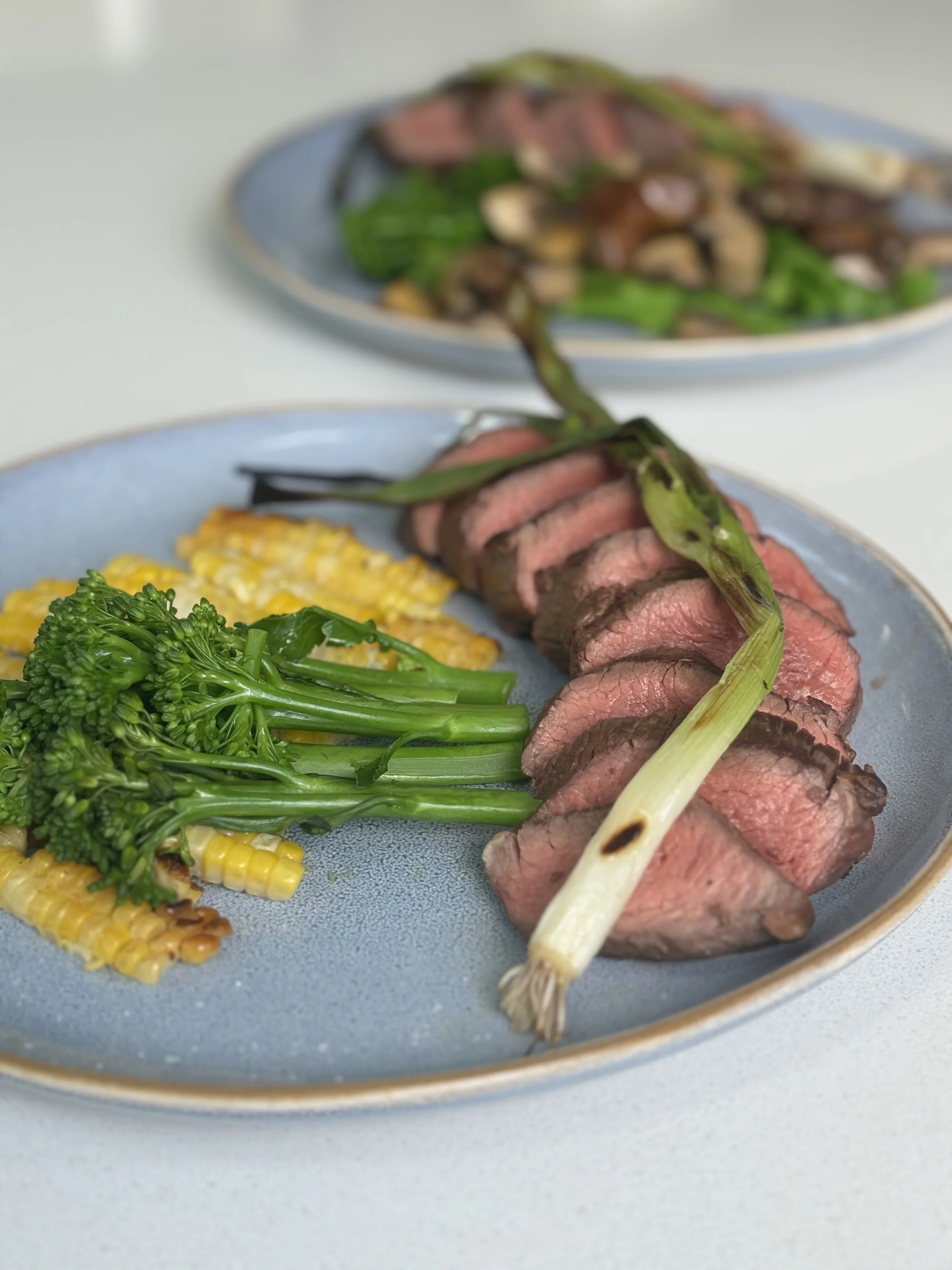 Sliced medium-rare roast beef garnished with a grilled green onion, served with steamed broccoli and corn on the cob on a blue plate.