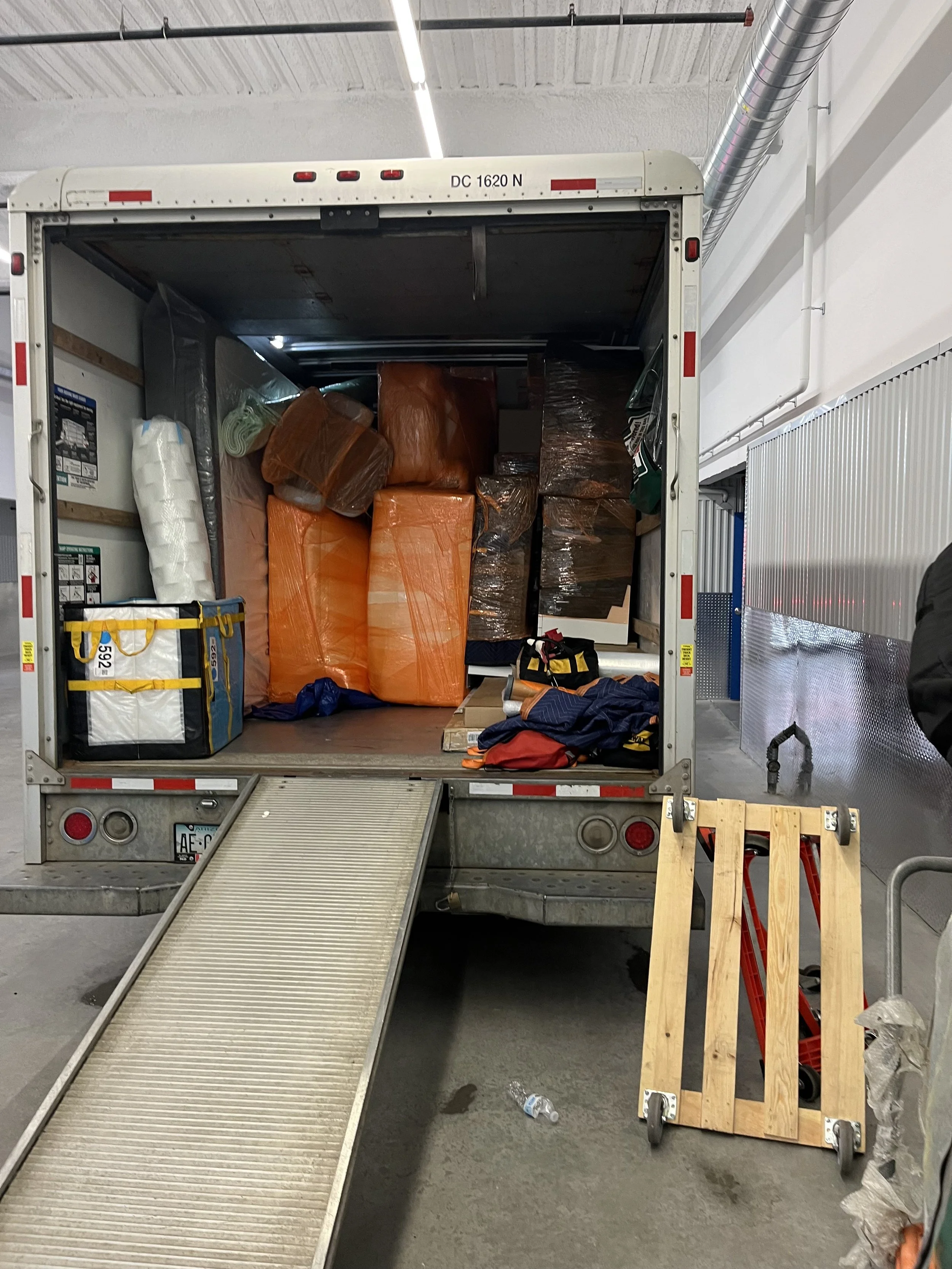 Open delivery truck with orange plastic bags inside, a ramp extends from the truck to the ground, and safety equipment is visible nearby.