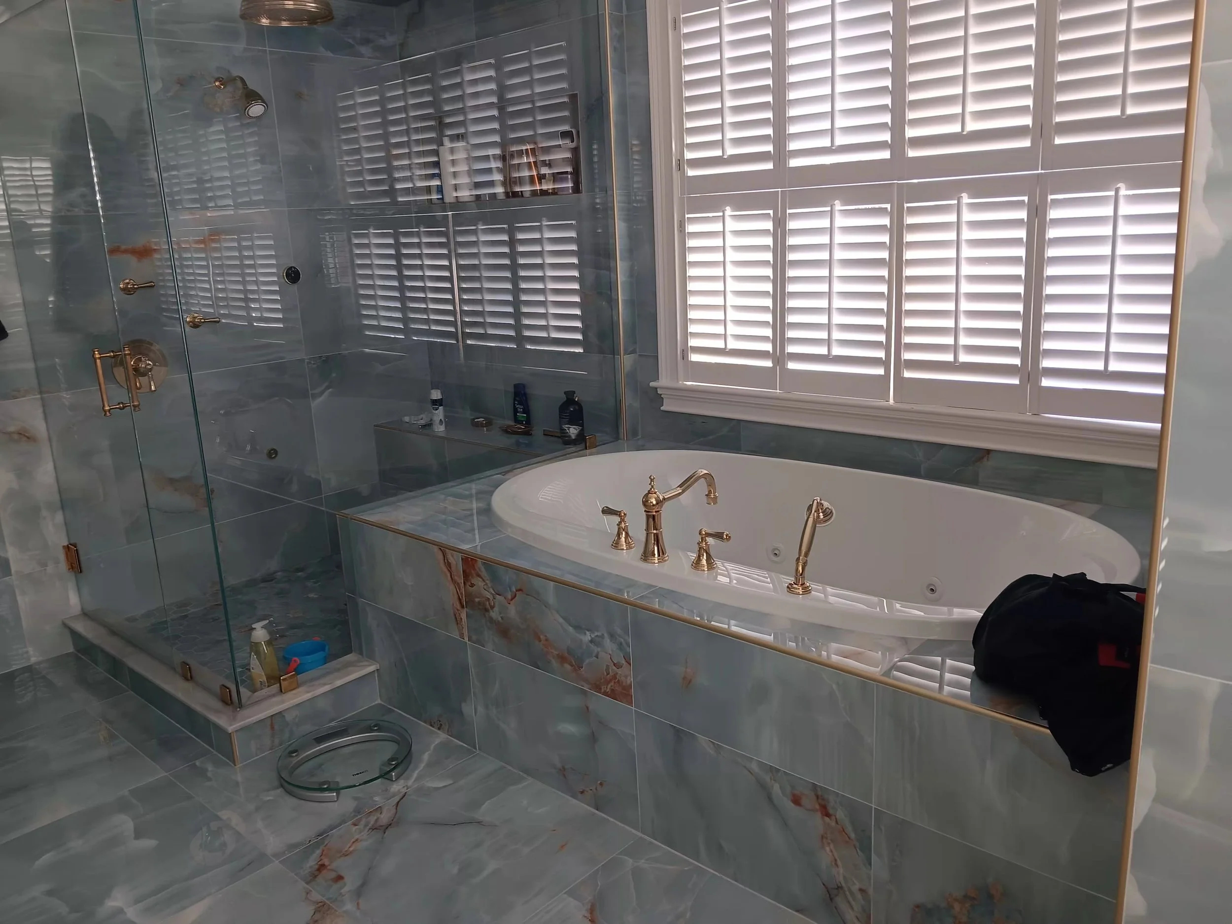 Modern bathroom with large window covered by white shutters, walk-in glass shower with gold fixtures, marble tiles, and a bathtub with gold fixtures. Bathroom items are on a small ledge and a digital scale is on the floor.