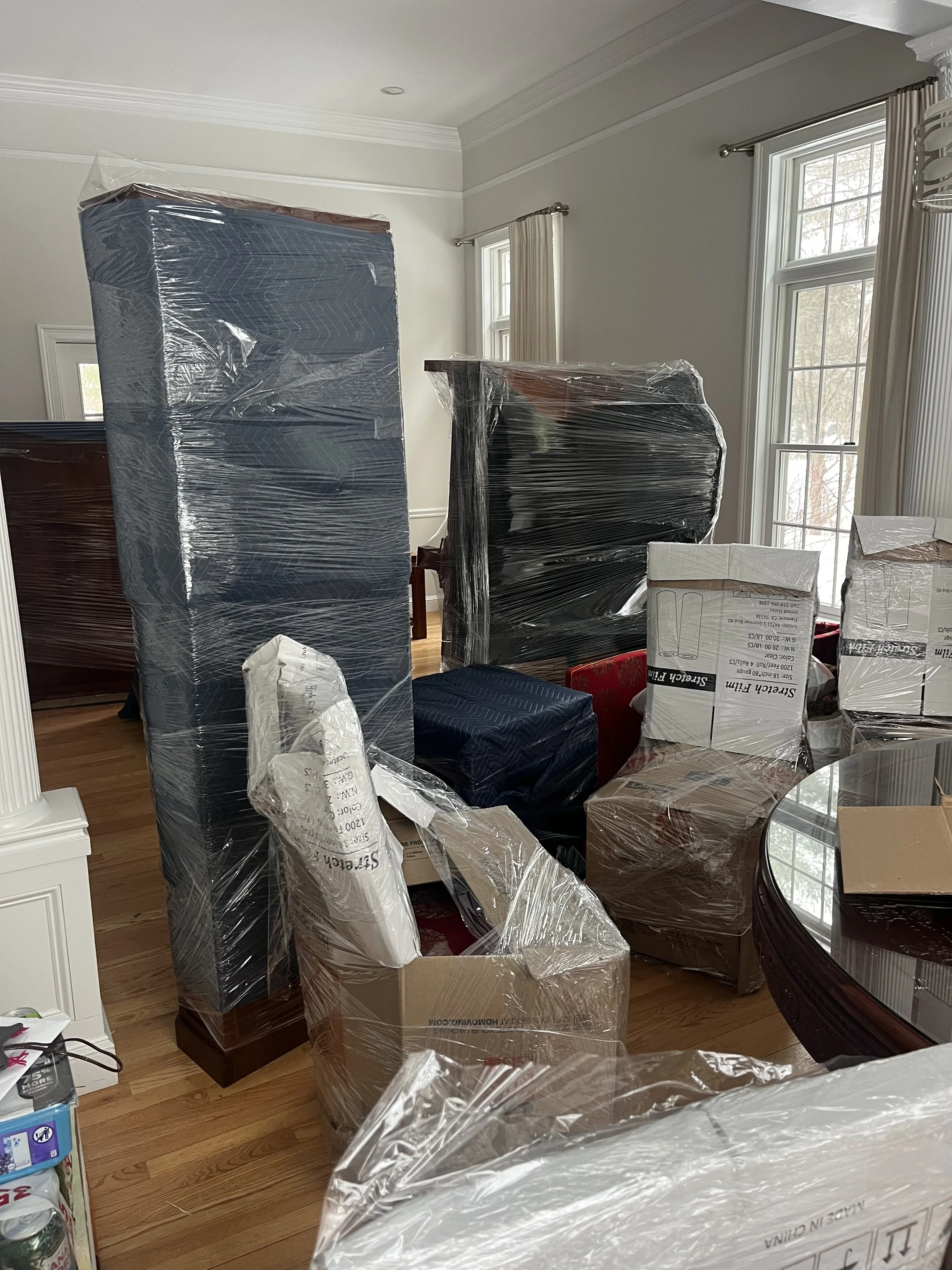 Moving boxes and furniture wrapped in plastic in a room with large windows and hardwood floors.