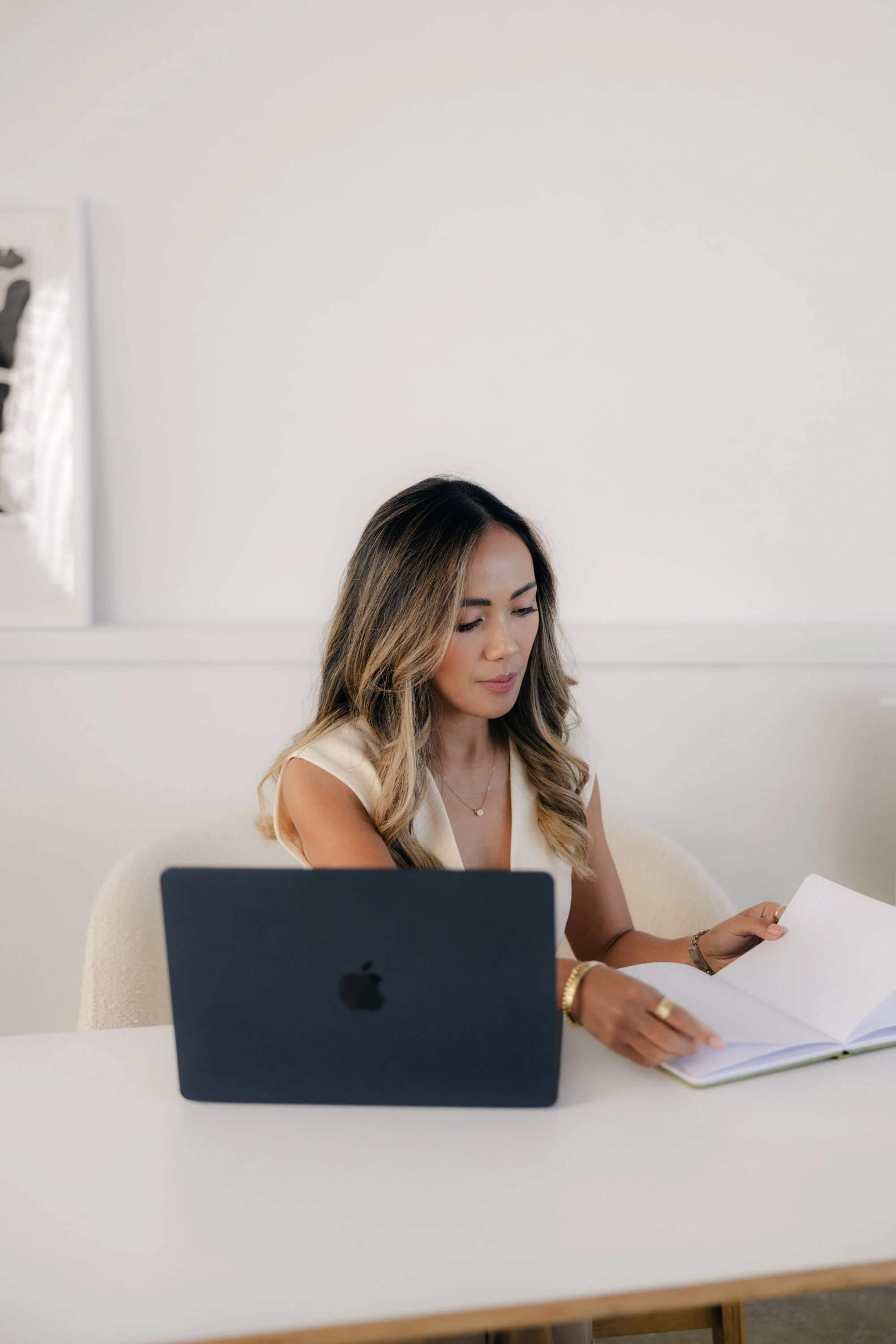 A woman with long wavy hair, wearing a sleeveless cream top, sitting at a white table in a modern office, reading a paper. A black computer monitor is on the table, and there is a white wall with a framed photo on the left side.
