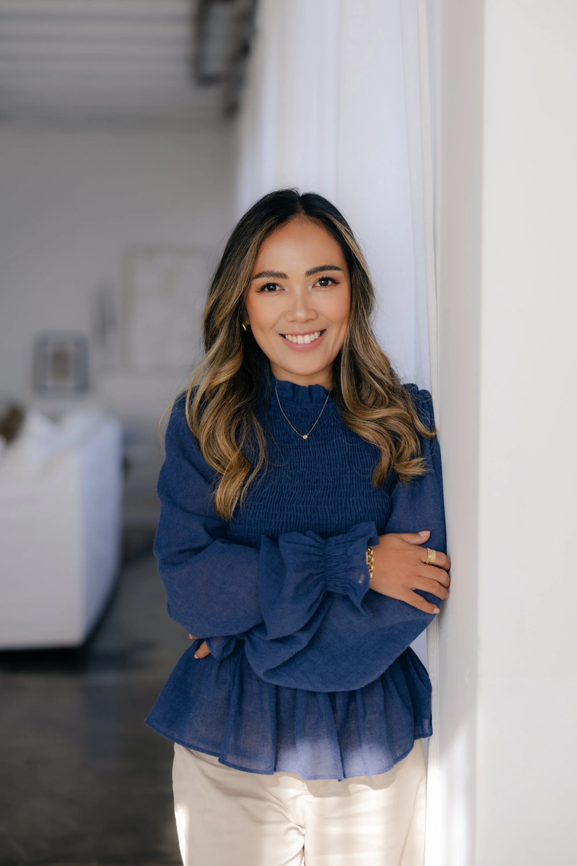 A woman smiling with crossed arms, wearing a blue top and white pants, standing in an indoor setting.