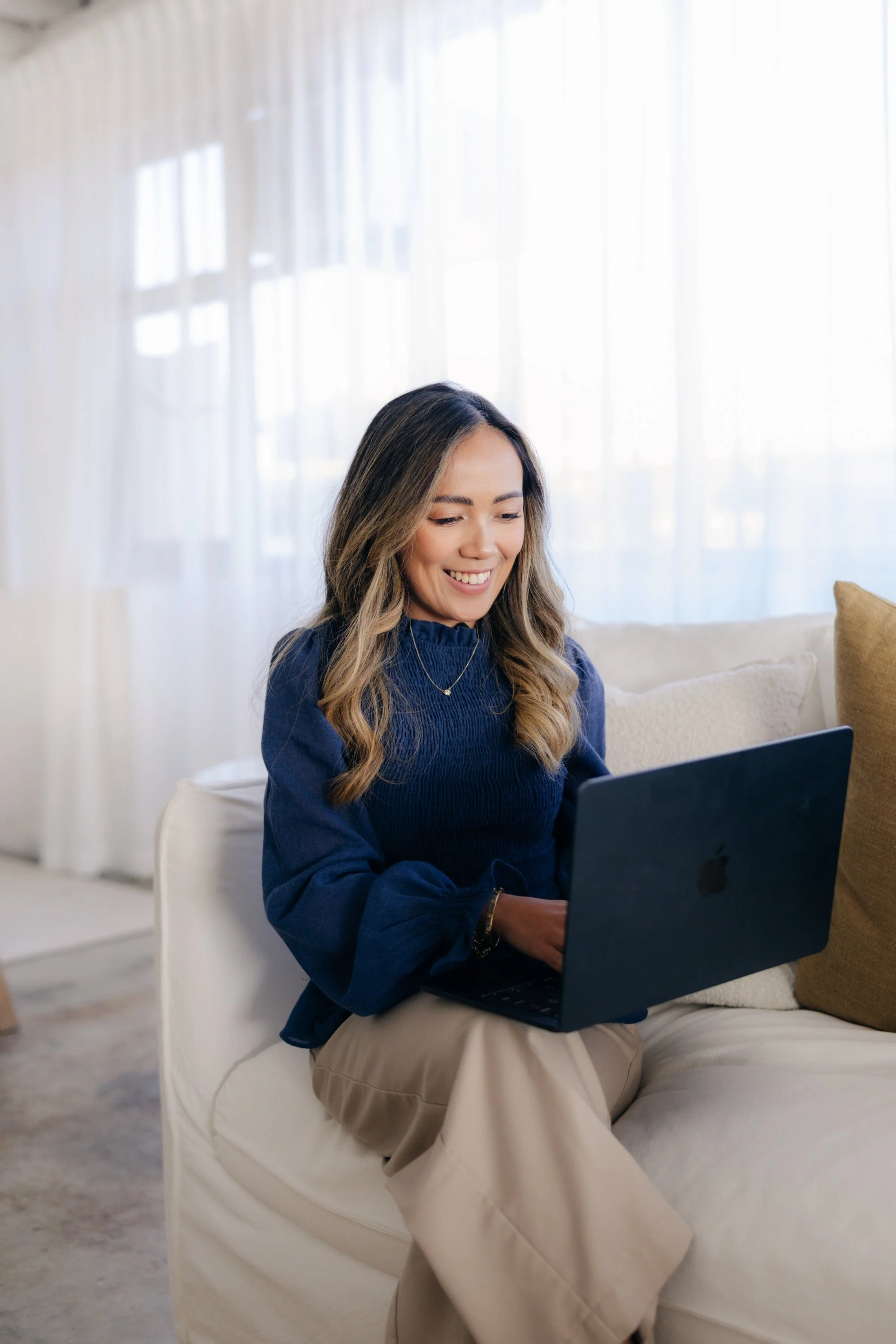 A woman sitting on a white couch, using a black laptop, with a beige cushion beside her, in a room with sheer white curtains and natural light.