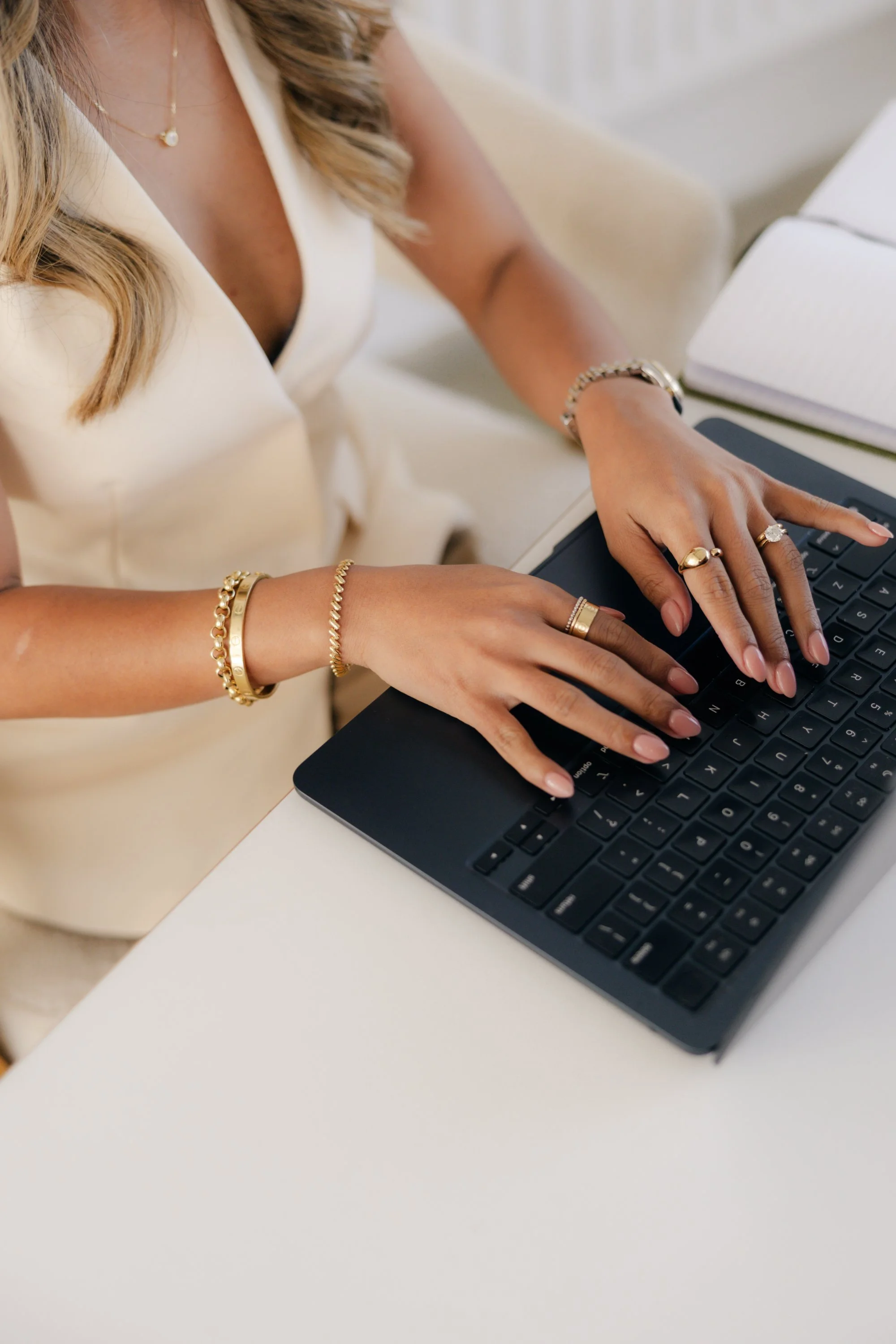 A woman wearing a cream sleeveless dress is typing on a black laptop at a white desk. She is wearing gold jewelry including bracelets, rings, and a necklace with a pearl pendant.
