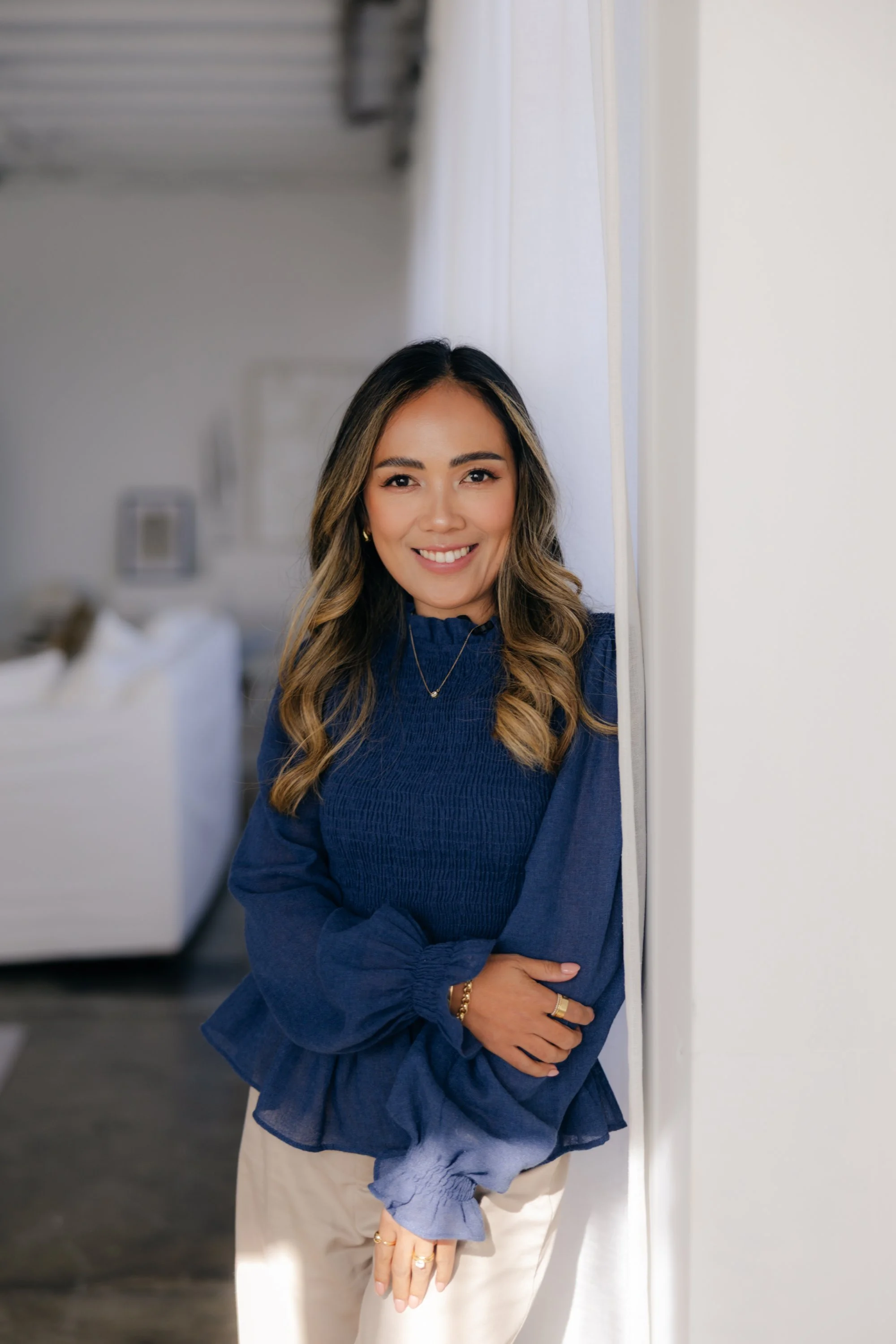 A woman with long, wavy hair and a warm smile standing indoors. She is wearing a dark blue, long-sleeved top and light-colored pants. She is leaning against a wall with her arms crossed in front of her.