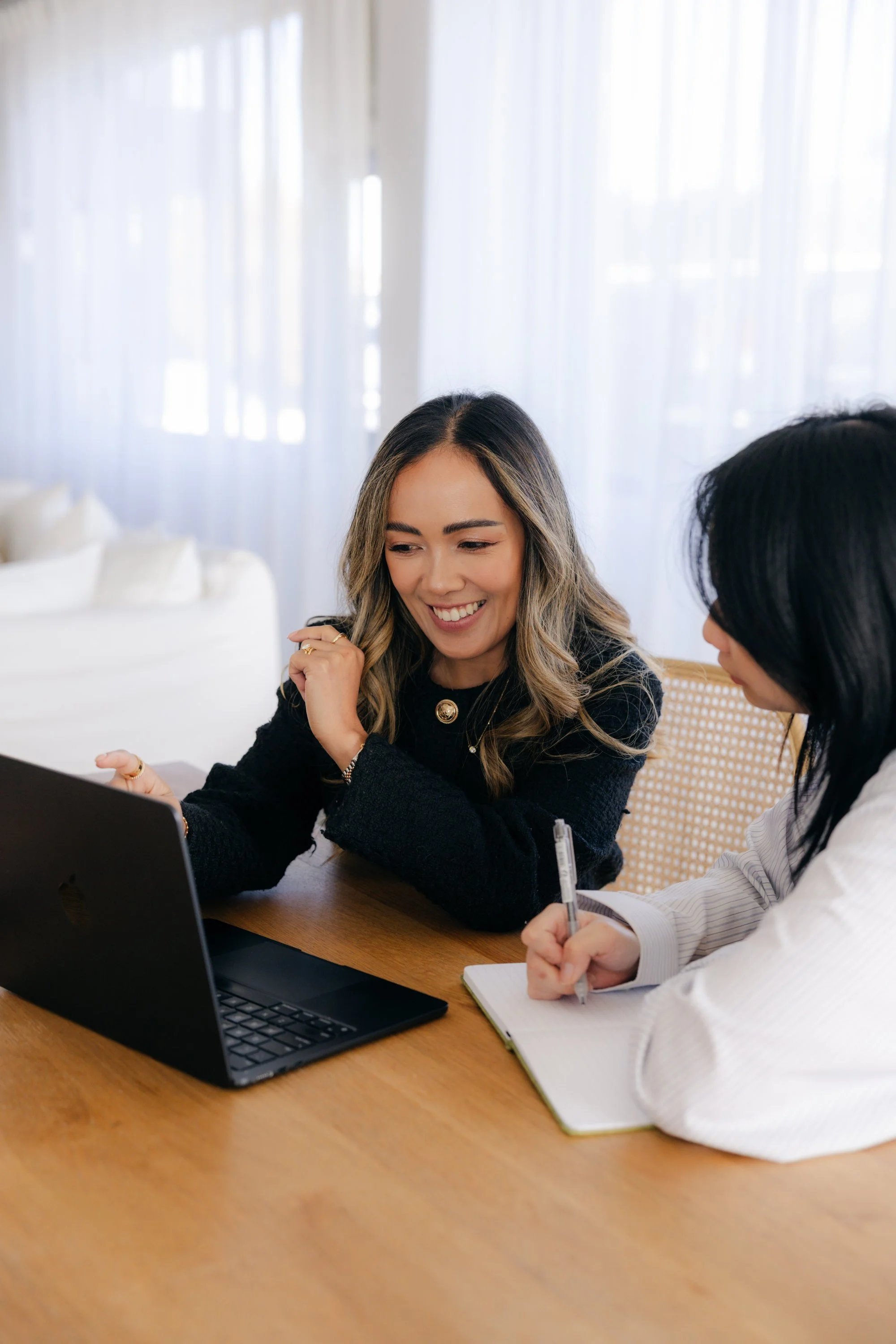 Two women sitting at a wooden table engaged in a conversation with a laptop, notebook, pen, and a notepad in a bright room with white curtains.