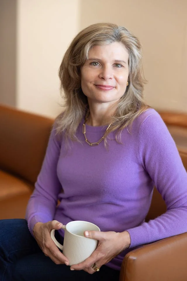 Maria Bertrand, IMFT, a female therapist seated on a leather couch in a purple sweater, holding a cup of coffee.