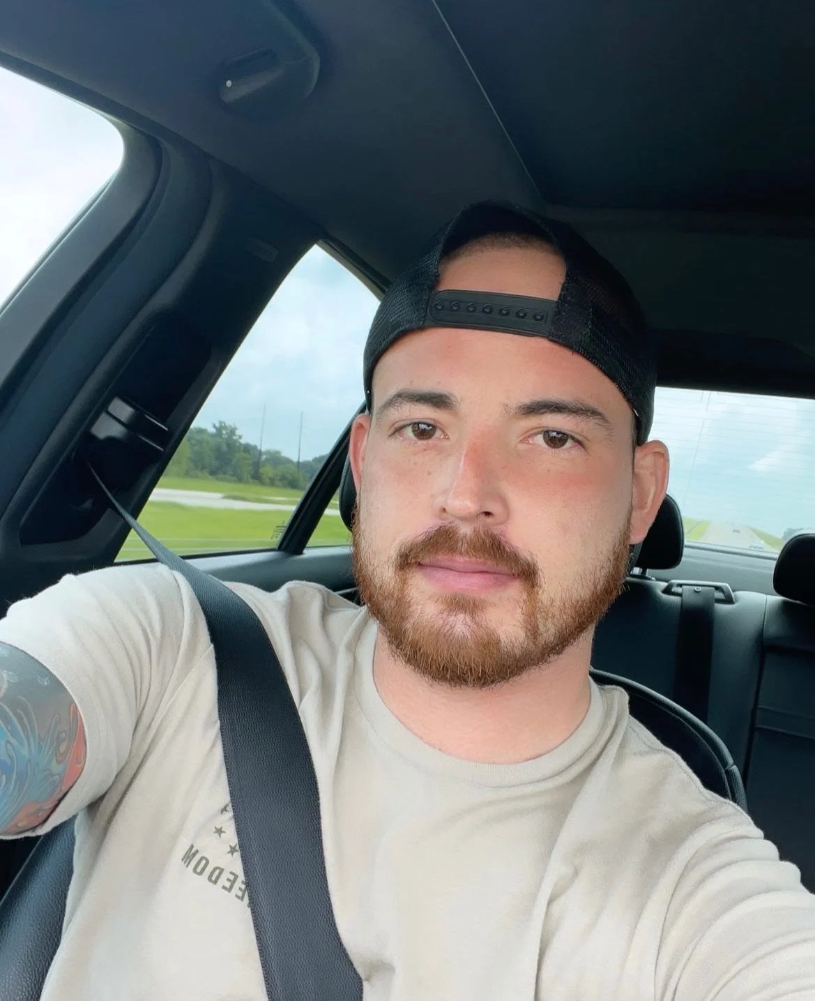 Selfie of a man with a beard and mustache, wearing a beige t-shirt and a black baseball cap backwards, sitting in a car with a seatbelt on, with a view of a highway and green landscape outside the window.