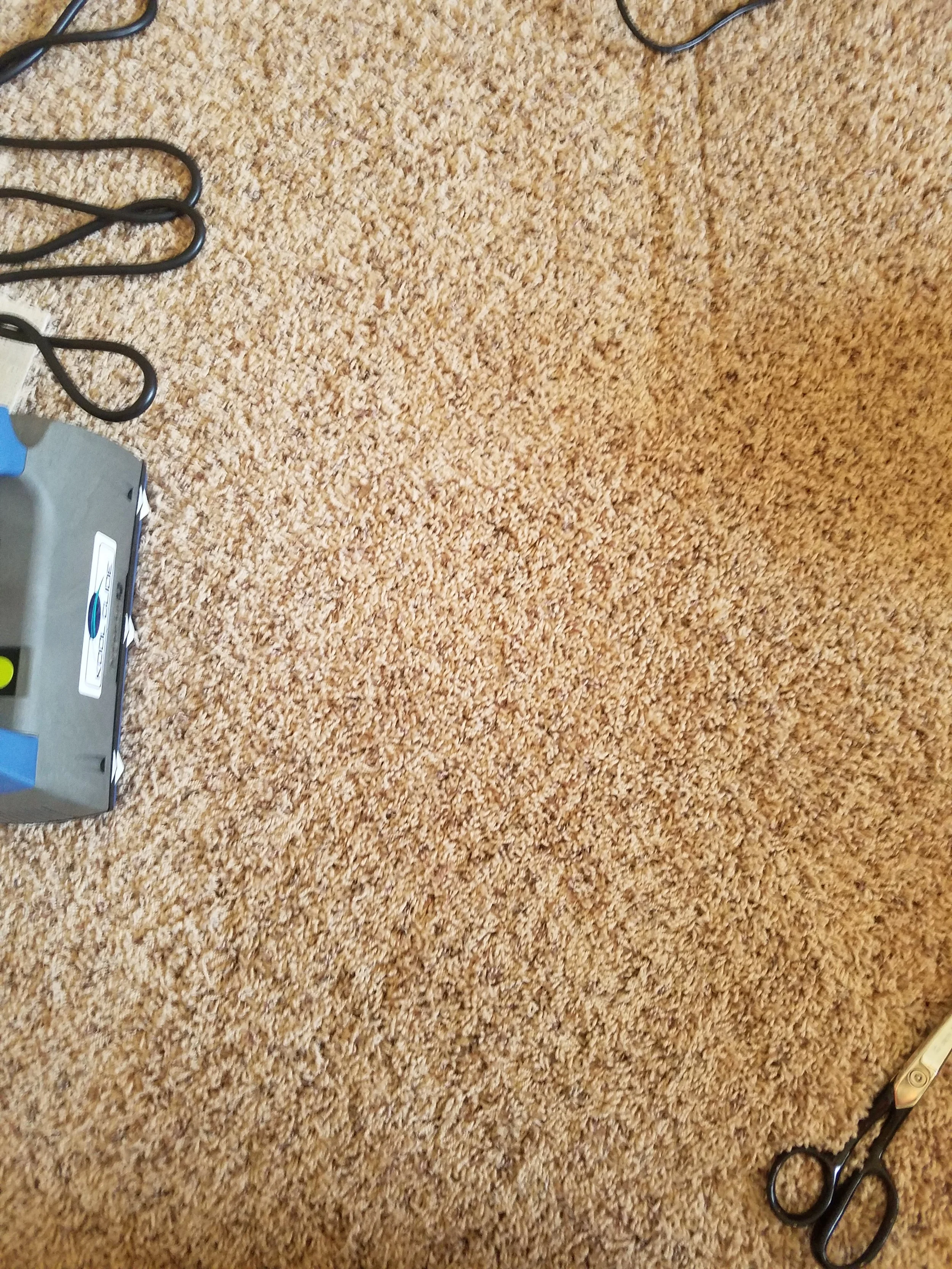 Close-up of a beige-carpeted floor with part of a gray device and black cords with loops on the edges.