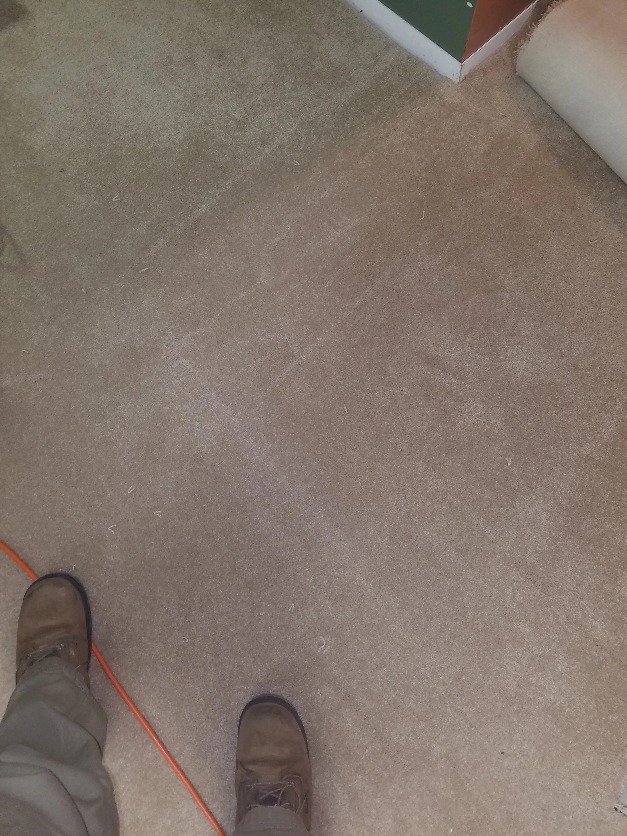 Lower perspective of a person standing on a beige carpeted floor, wearing brown boots and khaki pants, with some electrical cord on the floor.