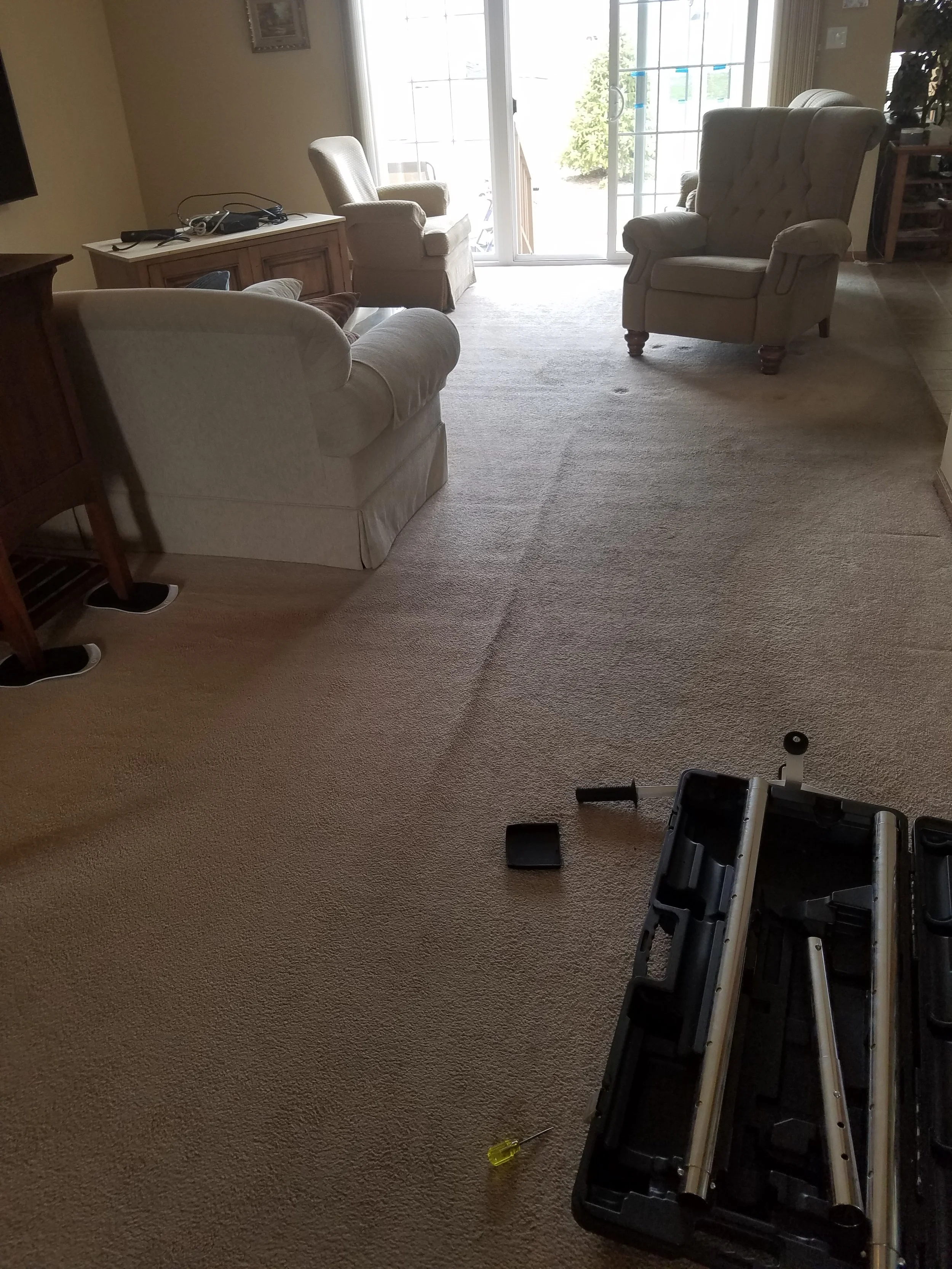 Living room with beige carpet, cream-colored upholstered chairs, a TV stand with electronic devices, sliding glass door leading outside, and a partially disassembled piece of furniture with a yellow screwdriver on the floor.