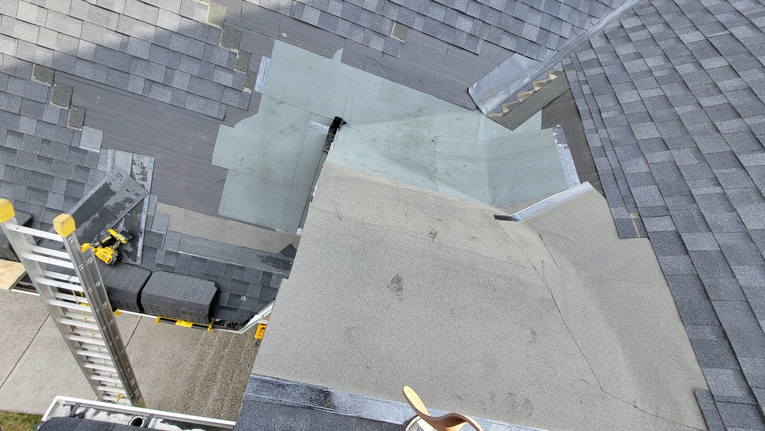 A rooftop with various shingles, decking, and tools for roofing work, including a ladder and cordless drill, with protective caps on the ladder's top.