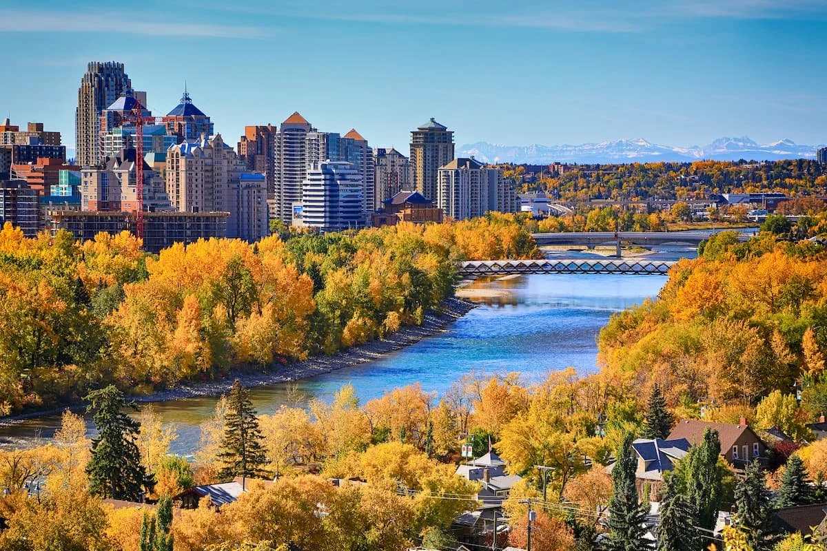 City skyline with skyscrapers and construction cranes, river with trees showing fall colors, houses, and distant mountains.