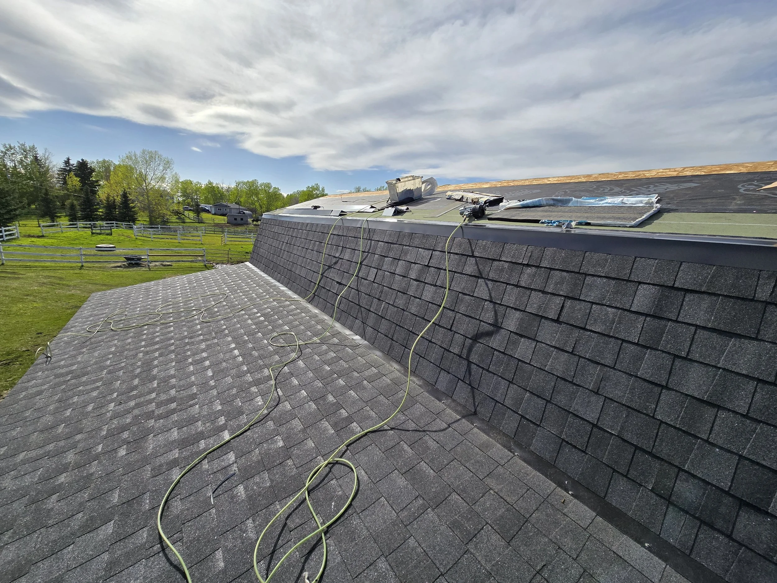 View of a rooftop with roofing materials and tools, and a cloudy sky, with trees and a fence in the background.