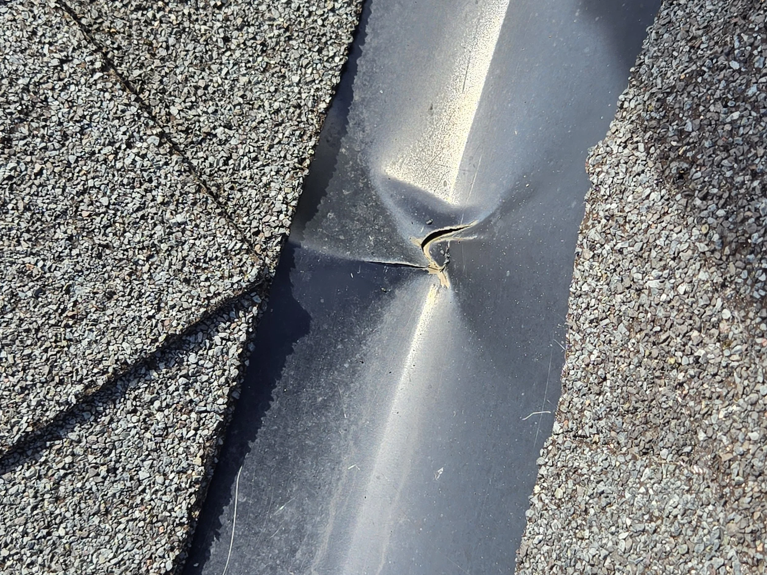 Close-up of a damaged metal roofing sheet with a tear, surrounded by gravel roofing material.