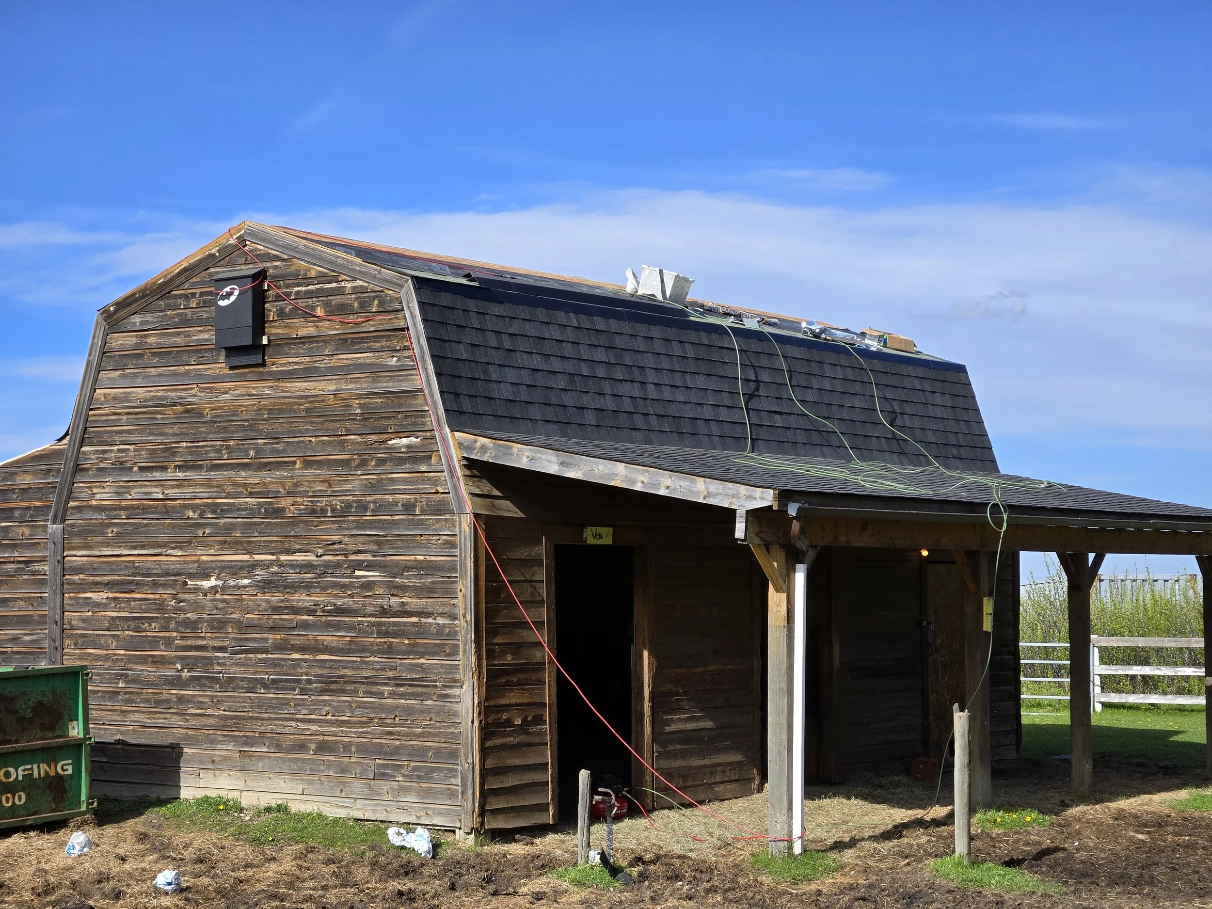 Wooden house under construction with solar panels on the roof and construction wires hanging, open side showing empty interior under a blue sky.