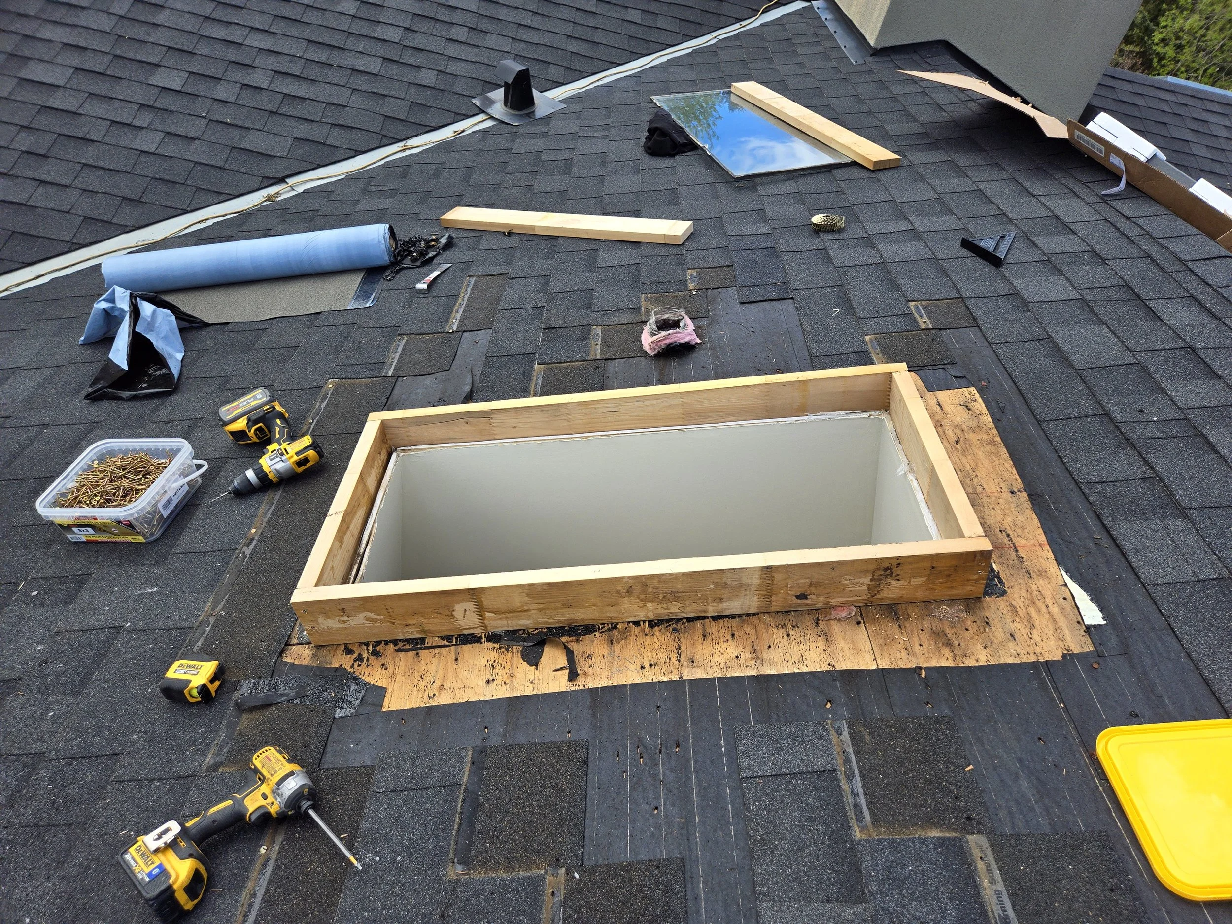 Roof with shingles under repair, open skylight frame, tools, and construction materials scattered around.