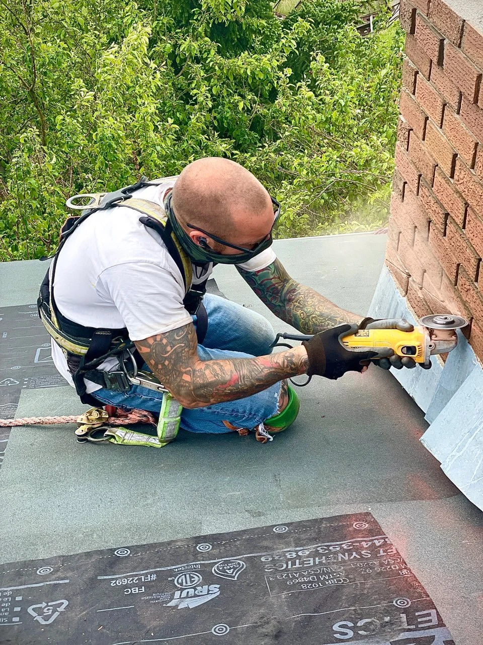 Shawn Turcotte the owner of turcotte roofing with tattoos, glasses, and a shaved head using a power tool on a brick wall on a roof.